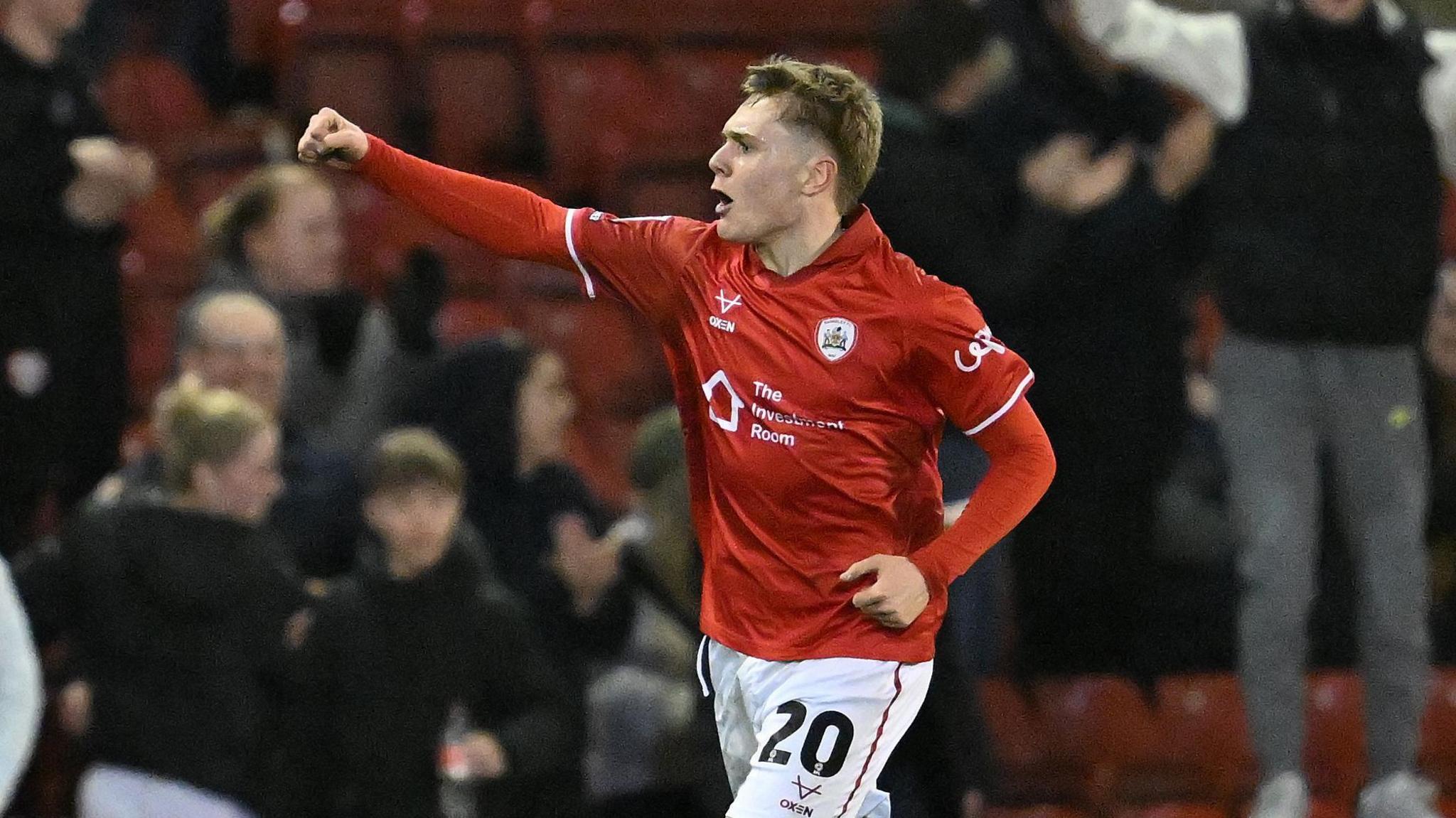 Charlie Lennon runs on the pitch with his right arm held in the air and fist clenched in celebration after scoring a goal. He is wearing Barnsley kit