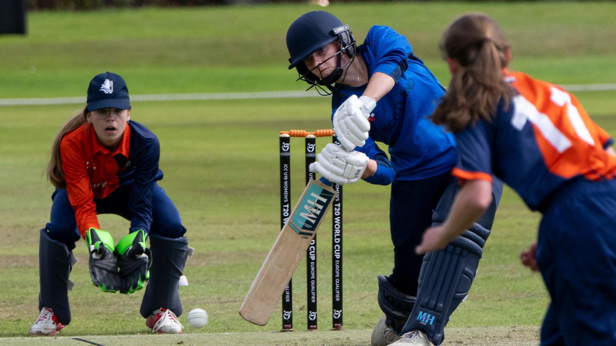 Niamh Muir in the blue cricket kit of Scotland swings her bat at a ball delivered by a long-haired woman in an orange and blue top while a wicket keeper in the same kit looks on hopefully