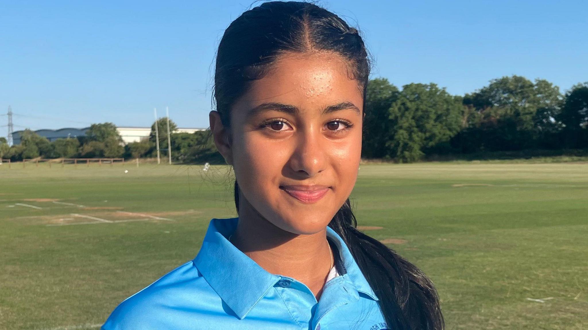 Anjni is standing on the short green grass of the cricket grounds and smiling at the camera. He has long dark hair in a pony tail and brown eyes. She is wearing a light blue cricket shirt with a collar and the logo for the Wellingborough Indians, which is a dark blue circle, almost like a wheel, with a star in the centre.
