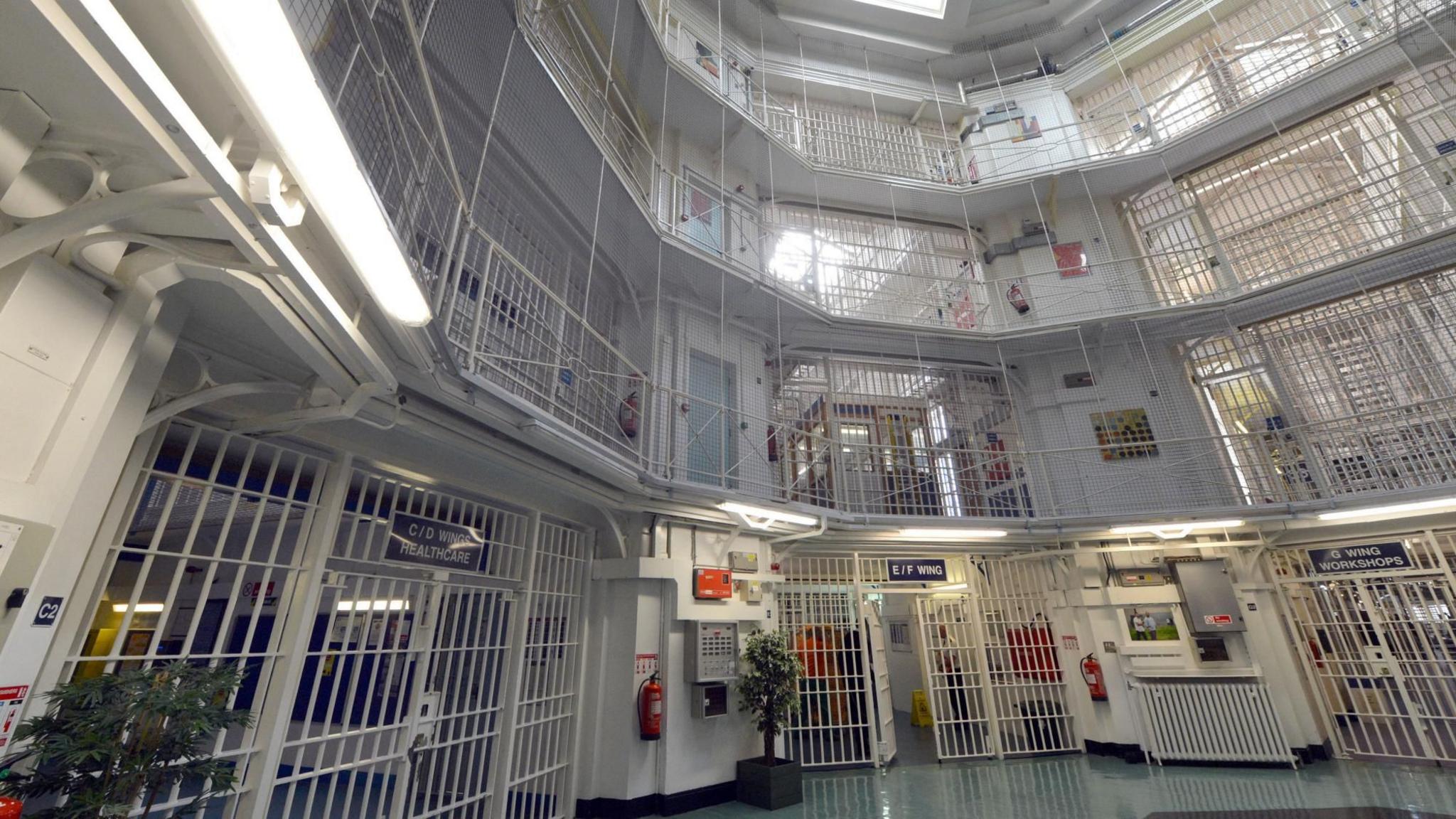 Interior of Pentonville Prison showing a multi-storey cell block with metal walkways, barred gates and protective mesh stretching up to a skylit roof.