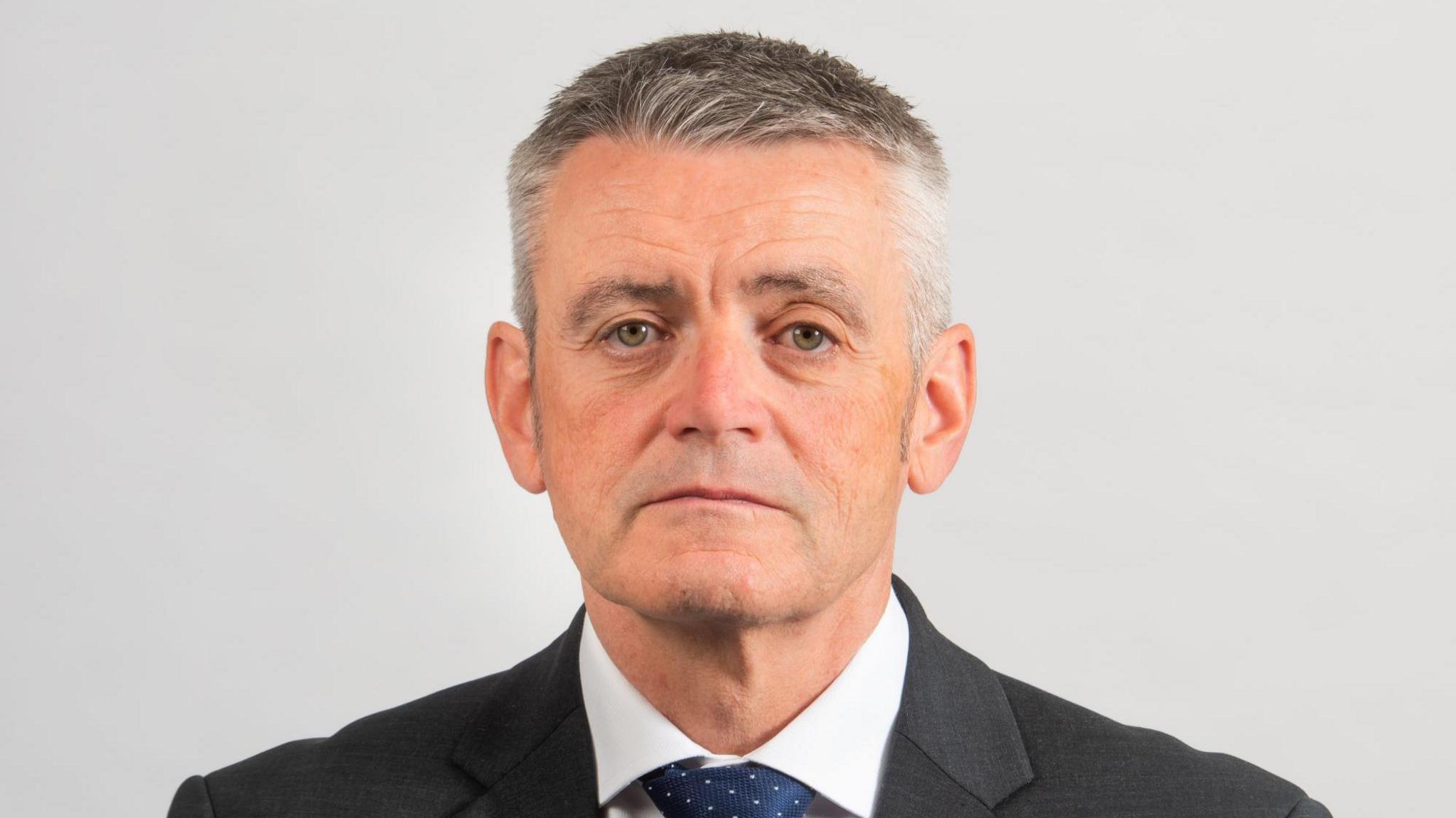 A man with short grey hair looks into the camera wearing a black suit with white shirt and navy tie. He sits in front of a white background.