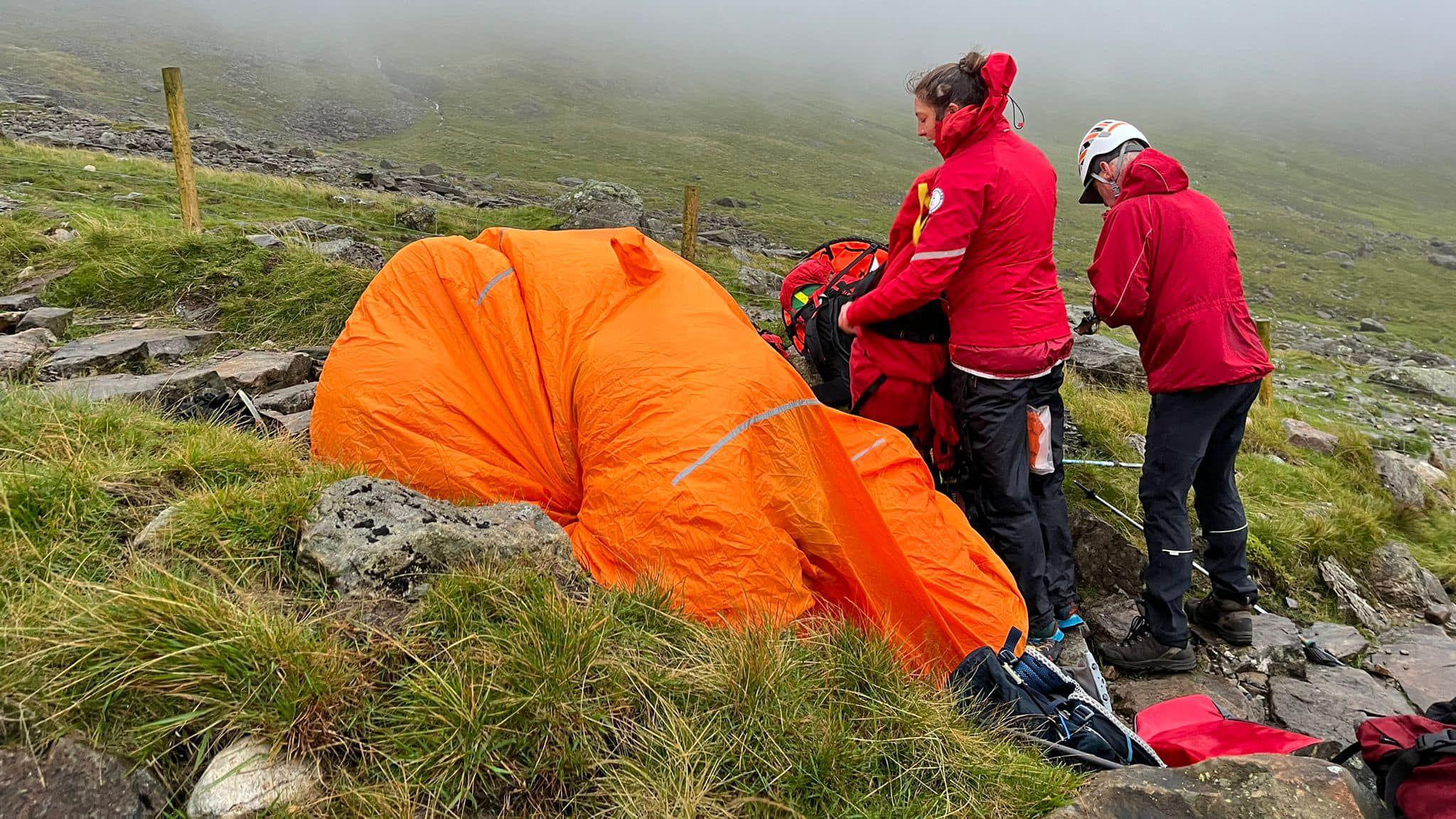 Mountain rescue volunteers approaching an orange group shelter with the casualty inside.