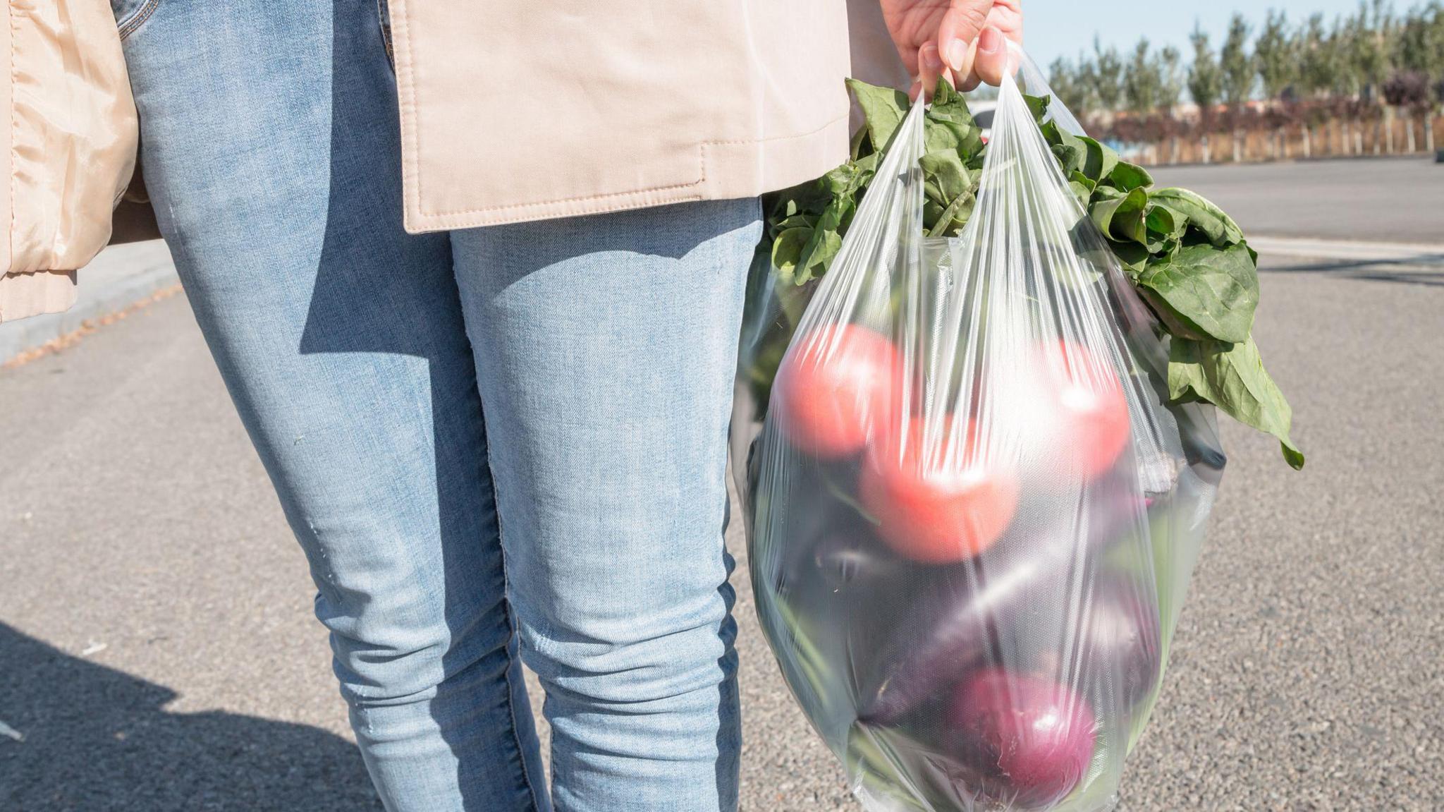 A woman holding a plastic bag of vegetables, including lettuce, tomatoes and aubergine. Only her bottom half is visible, clothed in blue skinny jeans, a grey t-shirt and beige raincoat.