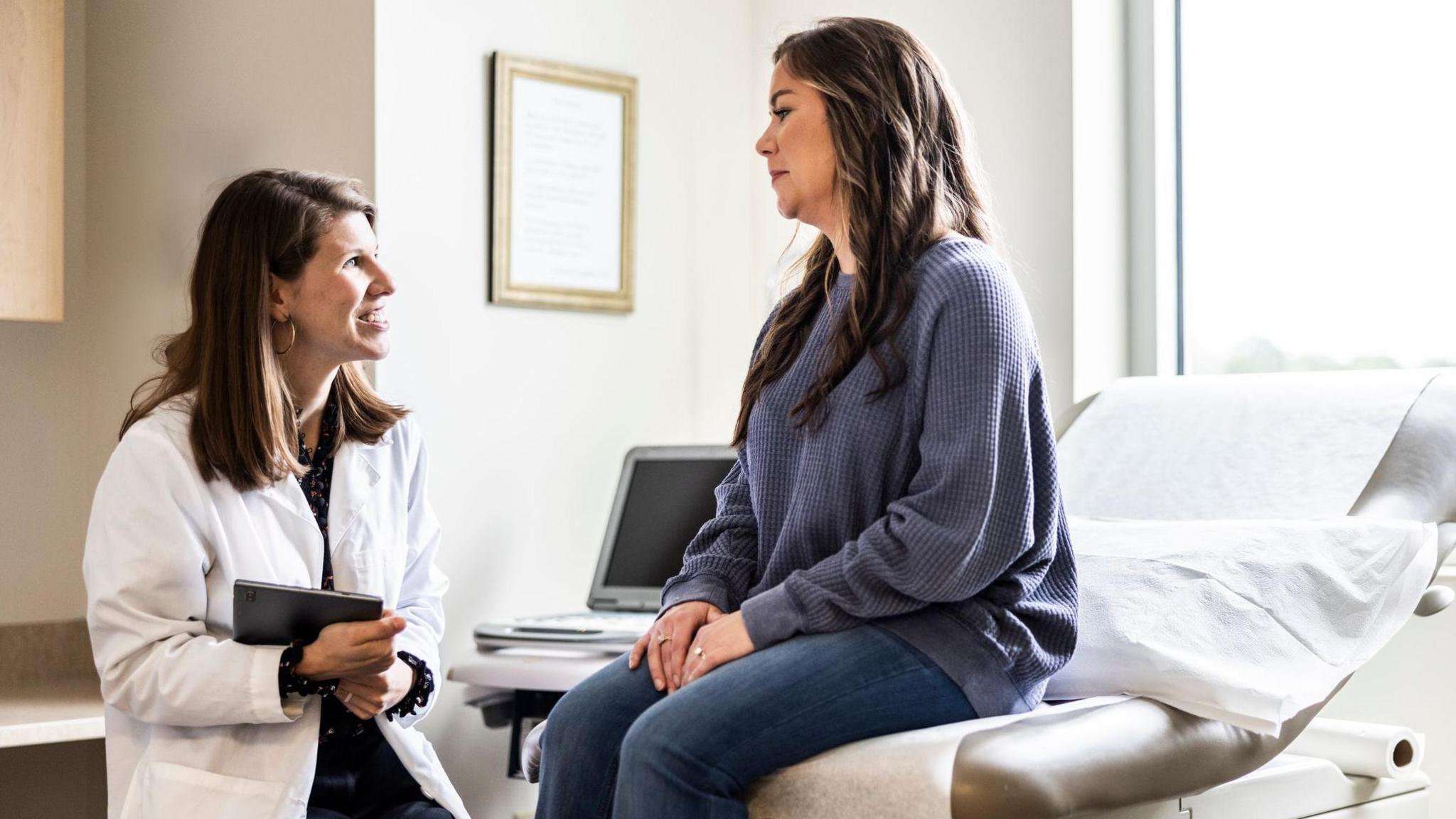 A Female doctor with mid length brown hair, wearing a white coat, holds an ipad and discusses treatment with female patient who sits on a bed, and wears a blue jumper in an exam room