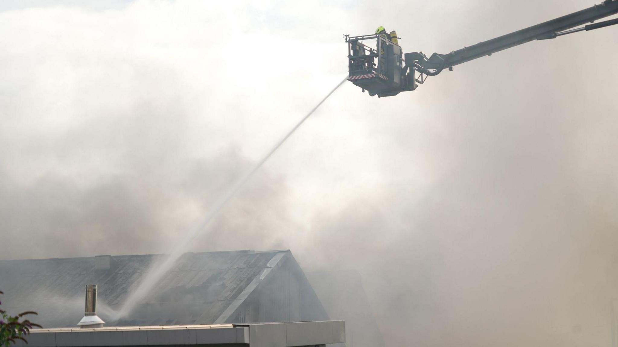A firefighter on a high reach appliance directs water towards a smoking building below.
