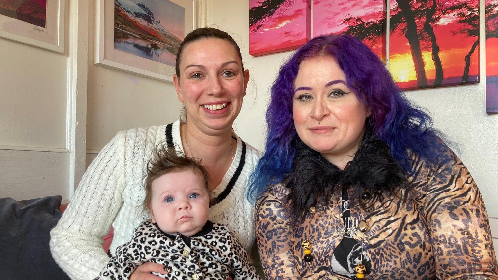 Frankie B wearing a white cable knit, V-necked jumper with a black band around the neck holding her baby daughter and comedian Maxine Wade who has purple/blue dyed hair and is wearing a leopard-print top with a black, furry colour. 