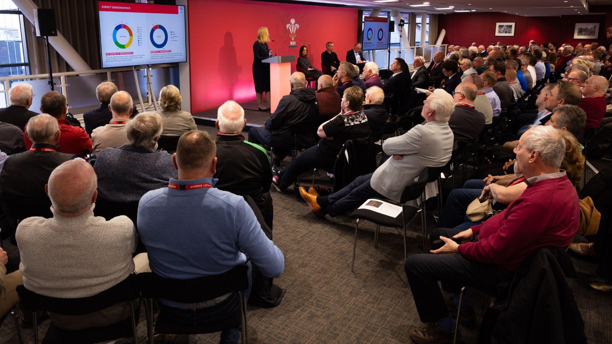Seated delegates at the EGM listen while Abi Tierney speaks at a lectern alongside other WRU officials on a low stage