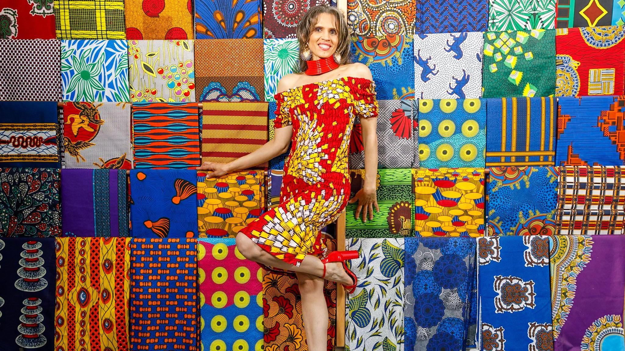 A woman with long brown hair stood in front of a multi-coloured quilted display. She is wearing an off-the-shoulder red dress, with a white and yellow graphic pattern, and red high heels. She has a thick red necklace and large earrings. She is posed with one knee raised and her hands touching the quilt.