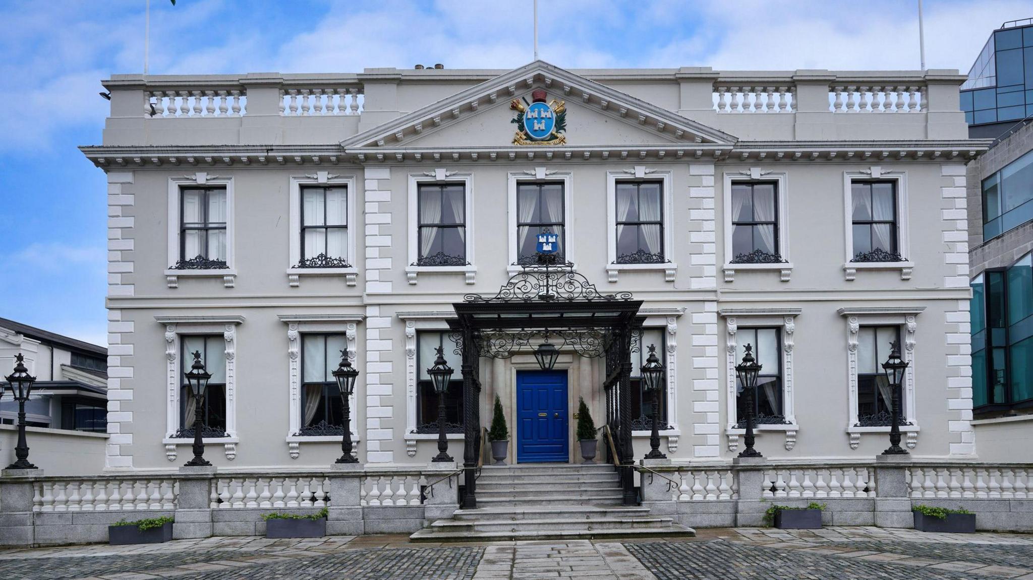 A grey brick building has steps leading up to a large blue door. It has seven windows on the first floor and six on the ground floor.