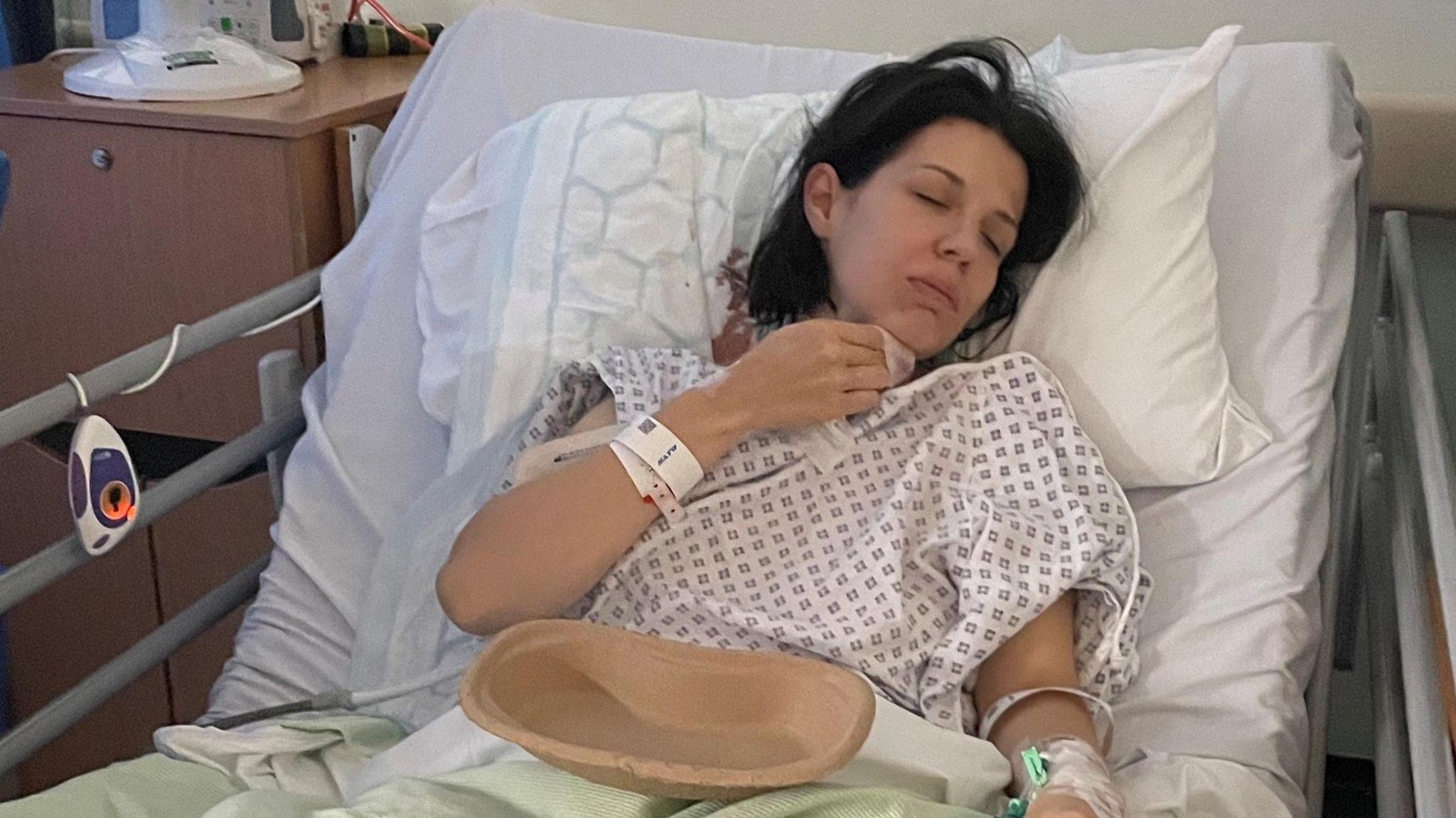 Mrs Griffin is lying in a hospital bed after an operation. Her eyes are shut and she holds a tissue near her face and has a brown bowl in her lap. She has medium length black hair and is wearing a white hospital gown and hospital wristbands.