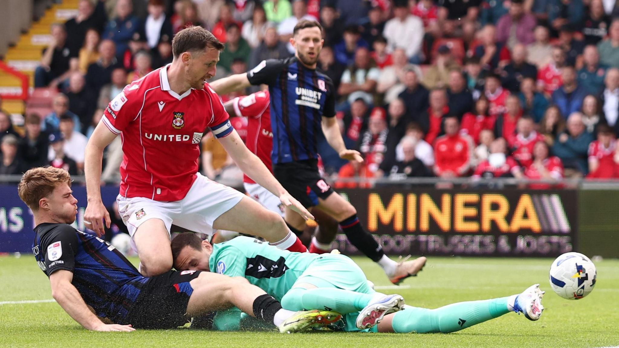 Action shot of a game between Middlesbrough, in red, against Wrexham in dark blue that shows a Wrexham defender and goalkeeper trying to stop the ball going into the net. 