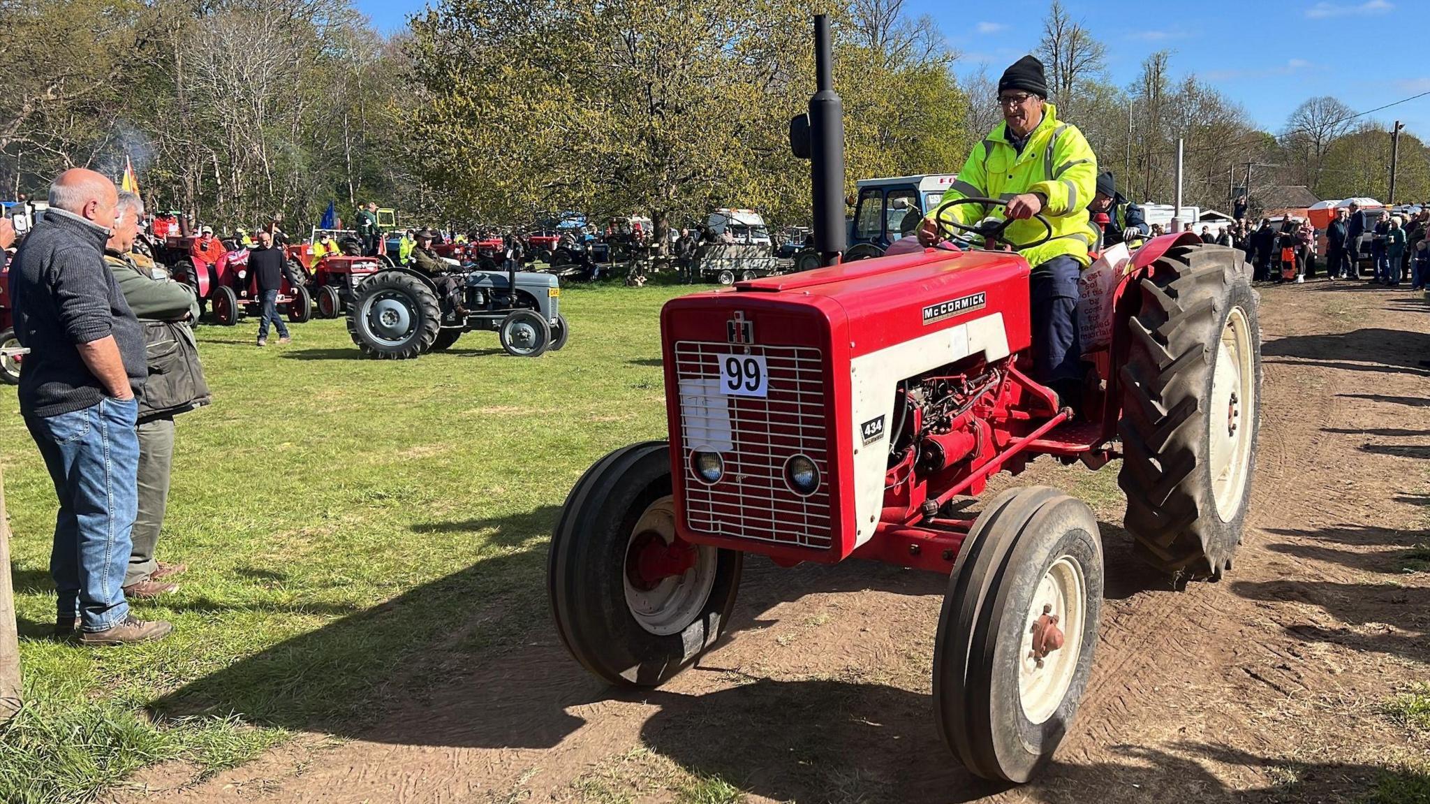 A man wearing a hi-vis jacket steers a red tractor along a dirt track as two people watch on. Lots of other tractors are parked in the background and being looked at by people.