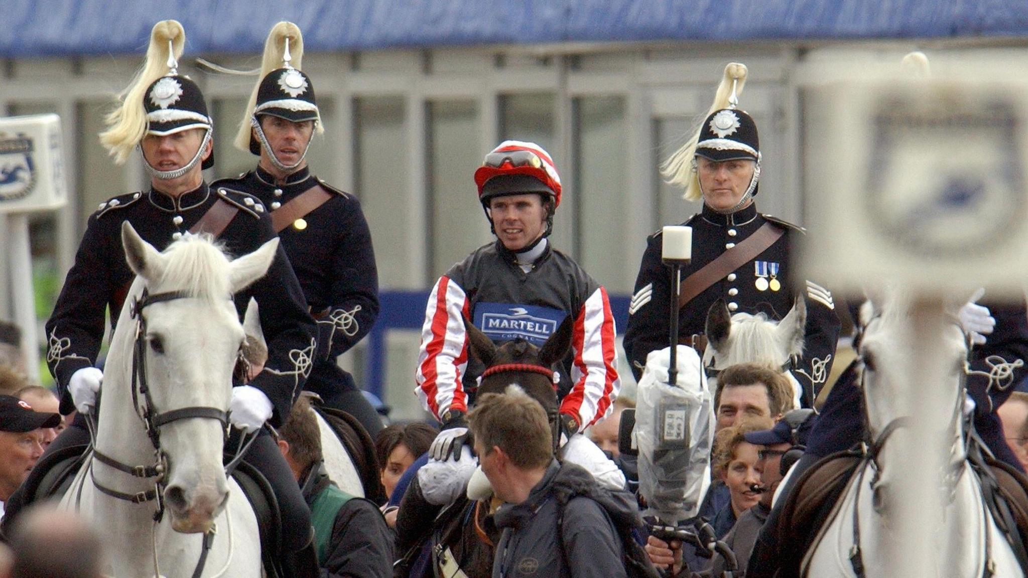Jockey Graham Lee at the end of the Grand National surrounded by people and police on horses. He is riding the horse Amberleigh House and is wearing colours which have a blue body and red and white striped sleeves.