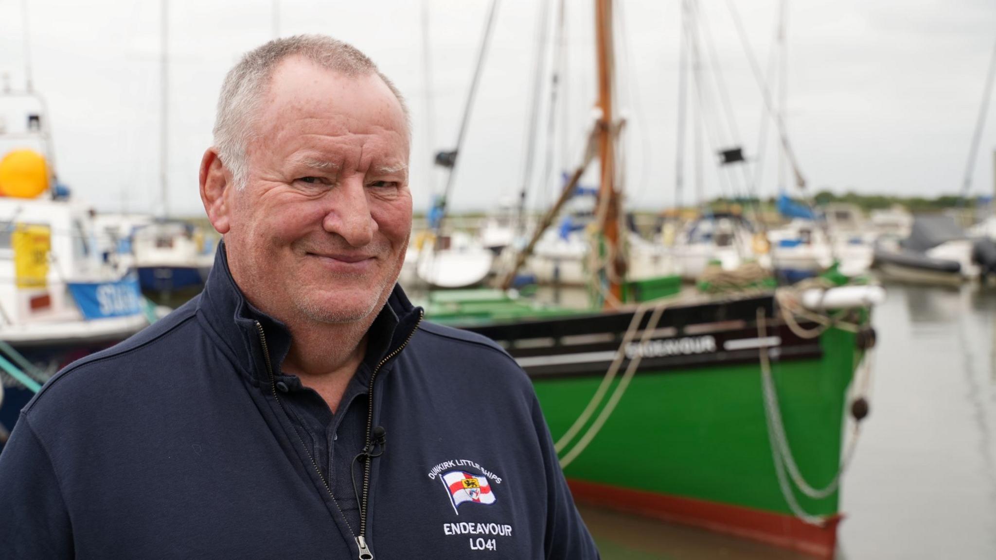 Cameron McGregor wearing a navy fleece standing on the decking at Leigh Marina, with the green Endeavour boat in the water behind him.