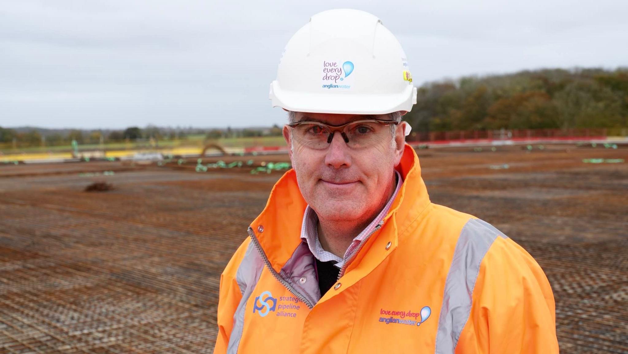 Andy Alder smiles at the camera while standing on a building site where a new pumping station is being built. He is wearing a white hard hat and clear protective glasses. He is also wearing an orange hi-vis coat. He has a small amount of grey stubble. 