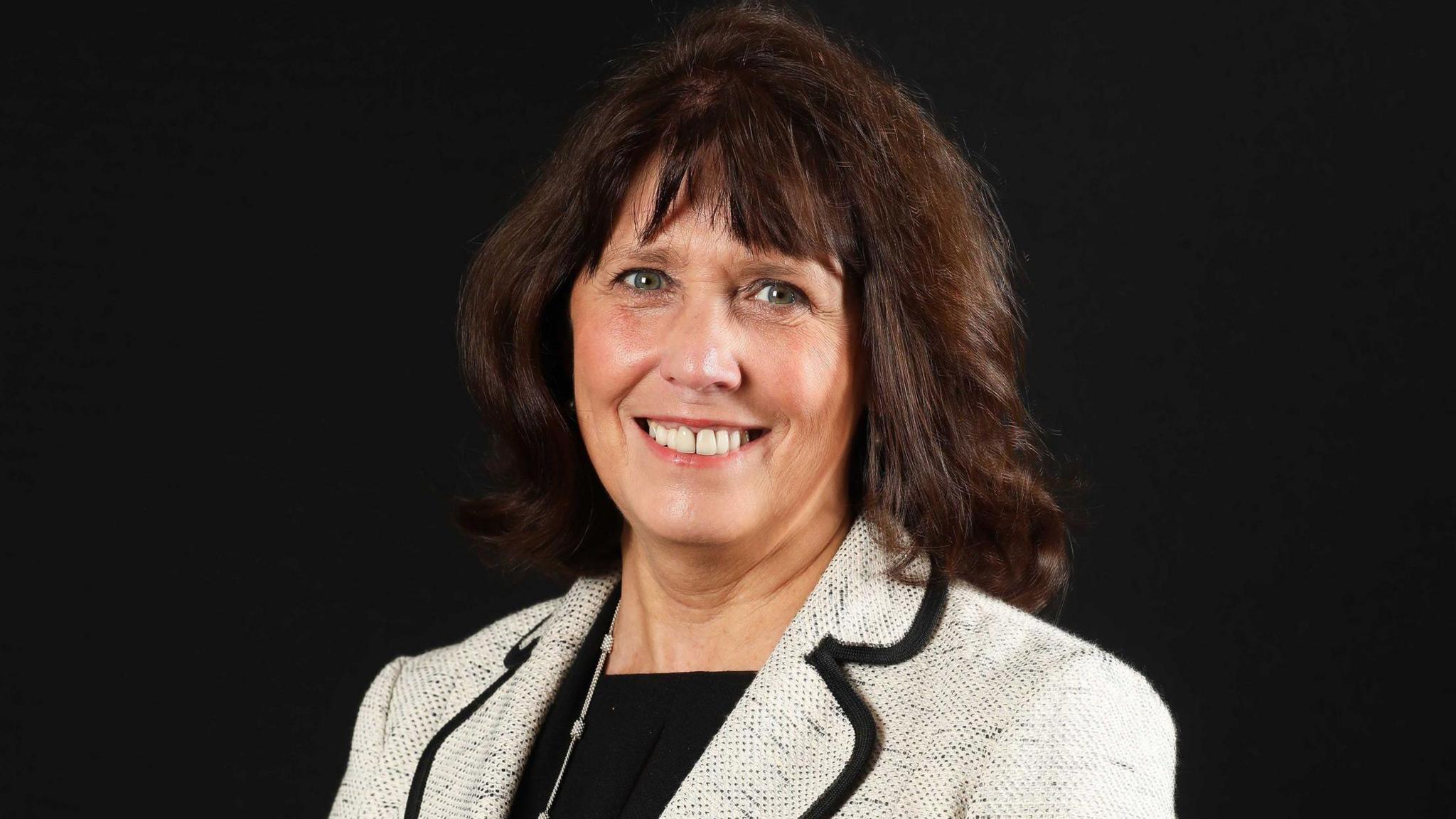 Ms Smith has shoulder length brown hair with a fringe and is smiling at the camera as she wears a cream coloured blazer with a black top. The background behind her is black.