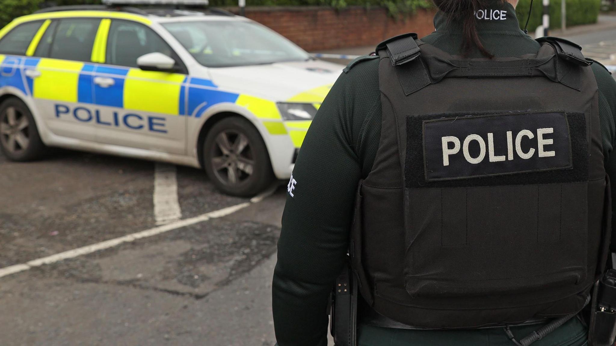 A police officer wearing a black bullet proof jacket and a green uniform with their back to the camera. A police car is parked next to the officer.