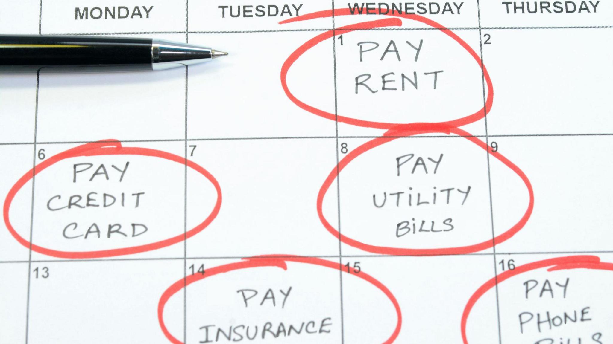 A calendar marked on different dates with 'pay rent', 'pay credit card', 'pay utility bills', 'pay insurance' and 'pay phone bills'. All are circled in red pen and a black pen also sits on the calendar. 