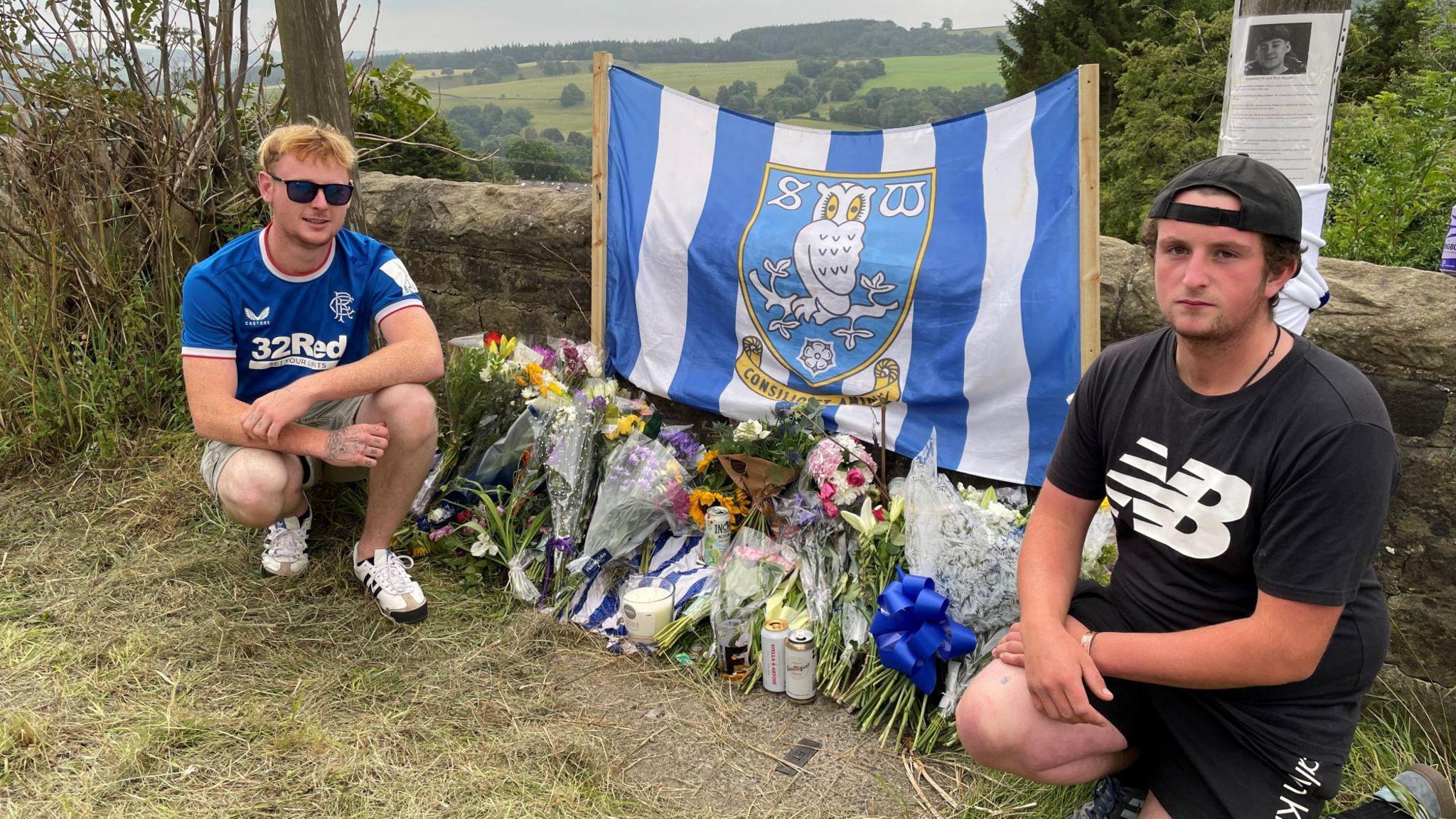 Two young men kneel next to floral tributes and a blue and white Sheffield Wednesday flag, which have been set up next to a stone wall.