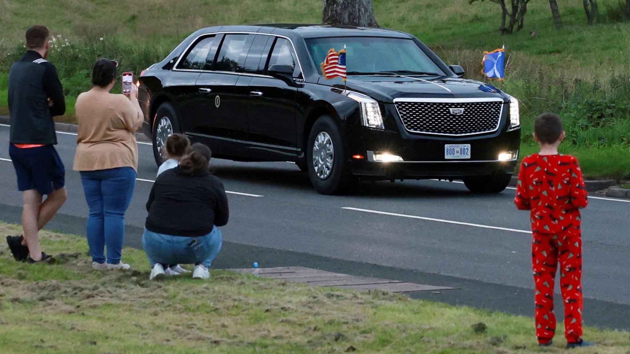 People take photos as the Trump motorcade passes by. There are three adults and two children watching, one of whom is wearing red pyjamas.