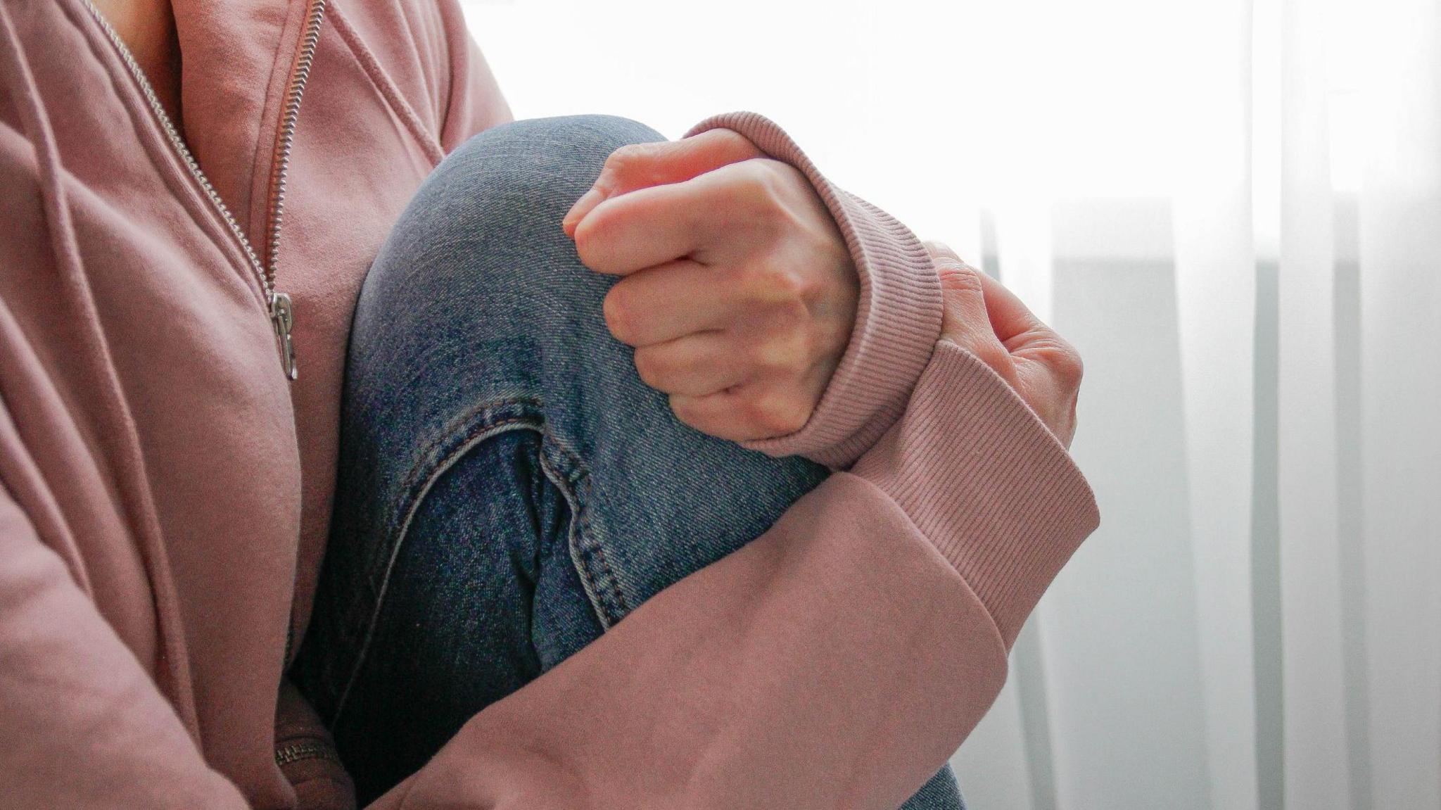 A person wearing a pink hooded top has their arms wrapped around their bent leg. They are wearing blue jeans and sat in from of a window.