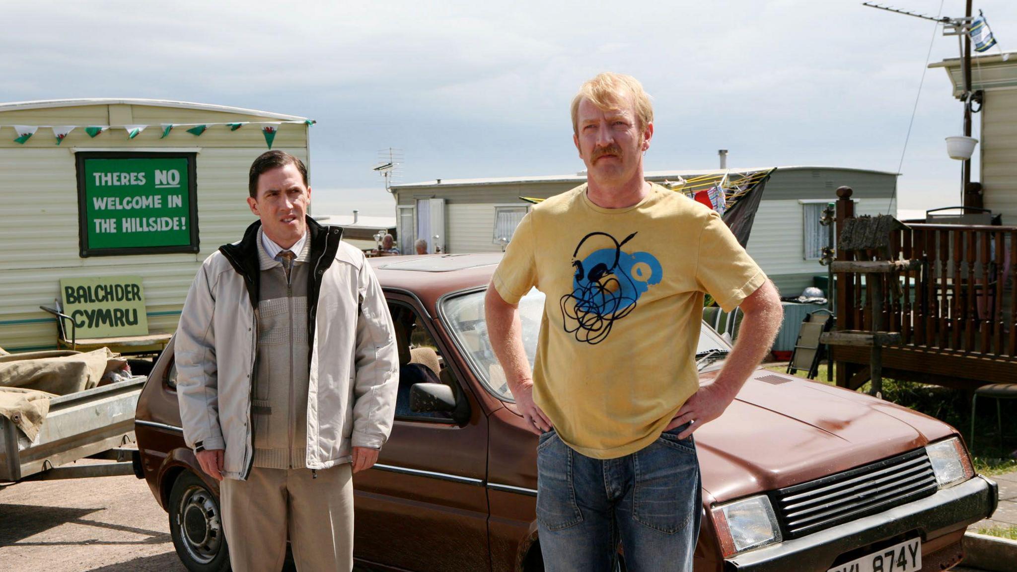 Two men stand in front of an old brown car at a caravan park