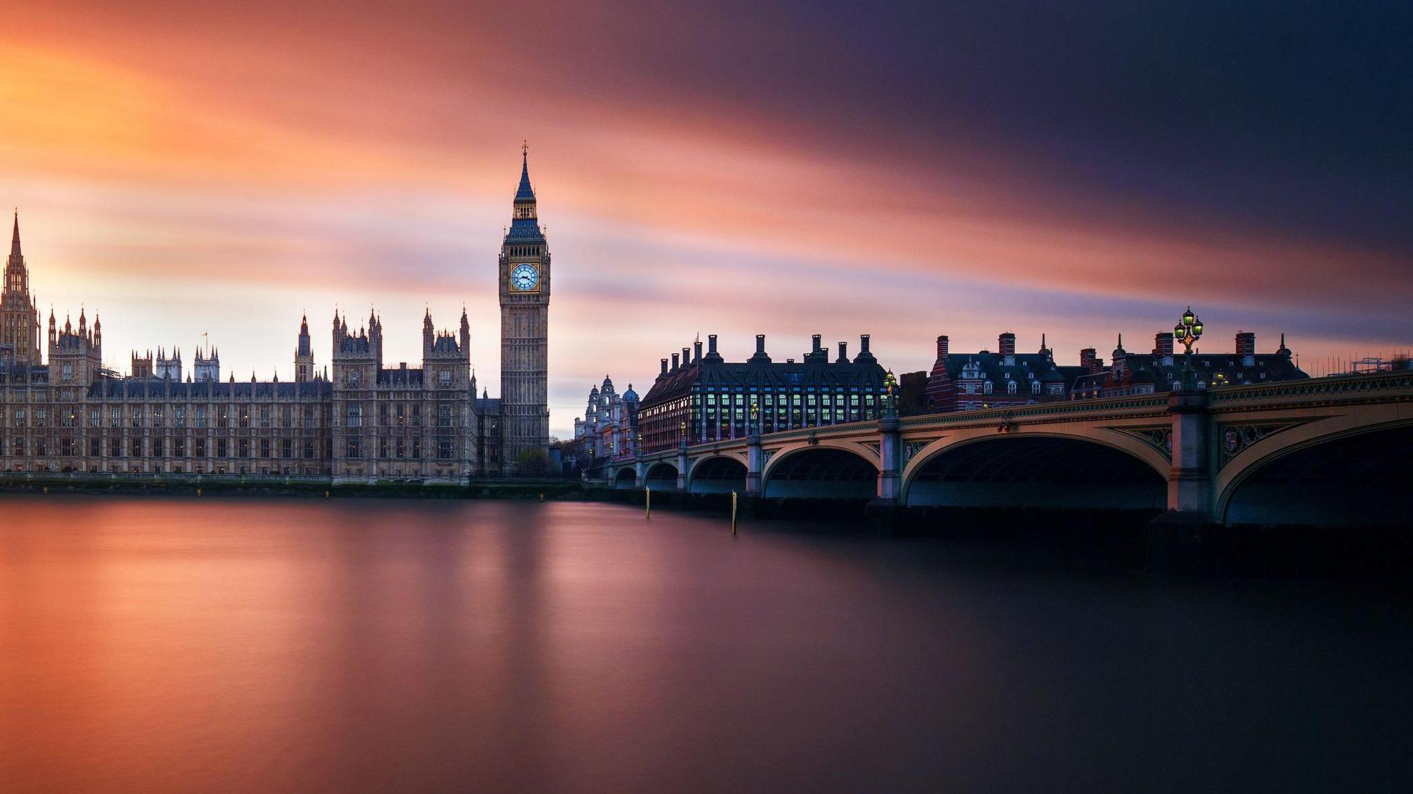 Ornate buildings and a tall clock tower on a river side with pink, orange and yellow sky reflected in the water below