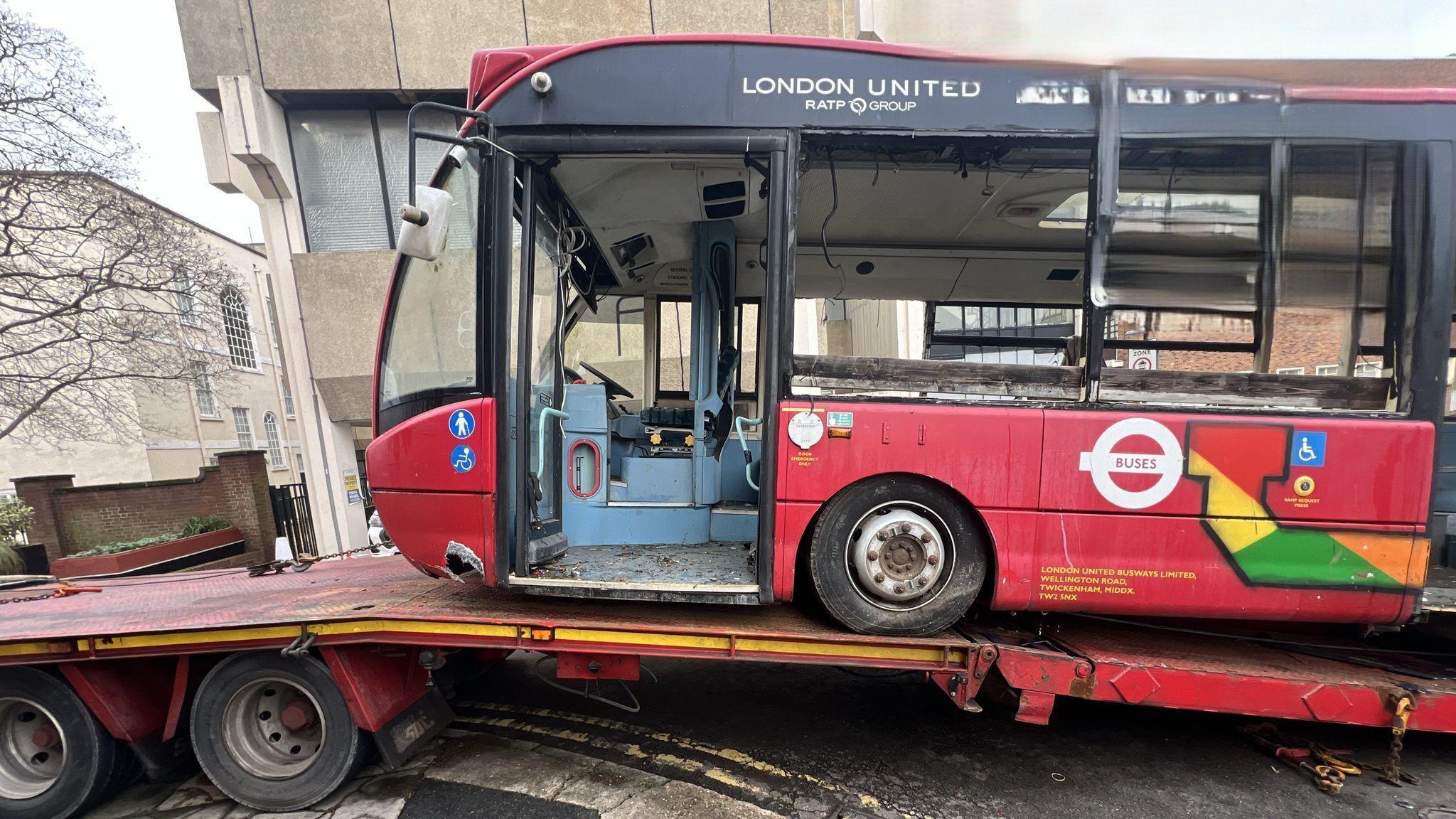 The side of the abandoned red bus, sitting on the bed of a truck as it is removed from the courtyard. The bus is a London United vehicle, with the door missing and all the windows smashed. The floor inside is filthy and the interior is painted a light blue. 