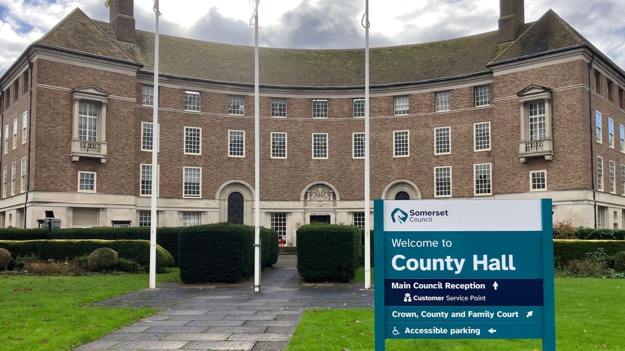 County Hall in Somerset. It is a brown brick building with lots of windows and flags outside. A turquoise sign reading welcome to County Hall is outside the building 