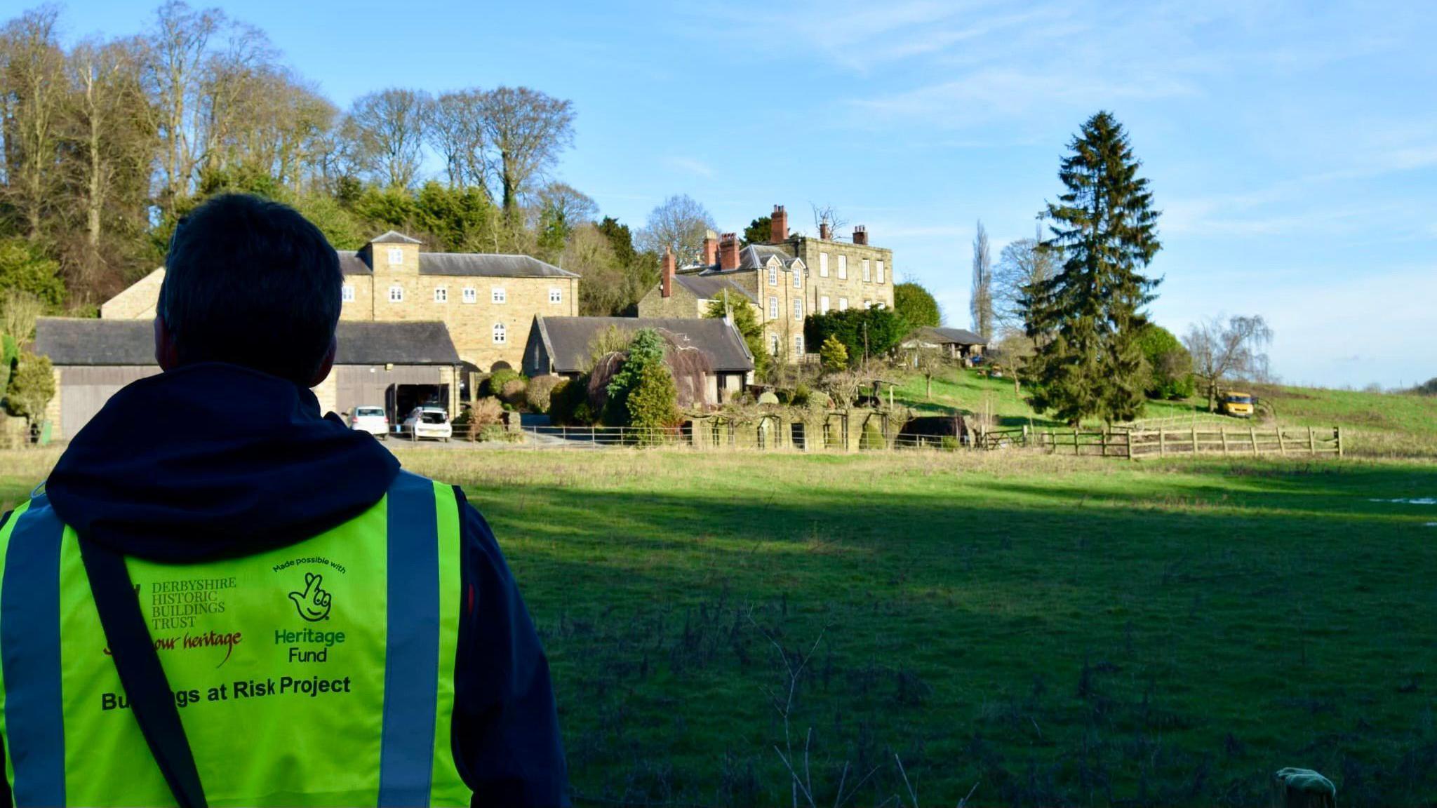 A man wearing a high-vis jacket looking across a field at buildings. 