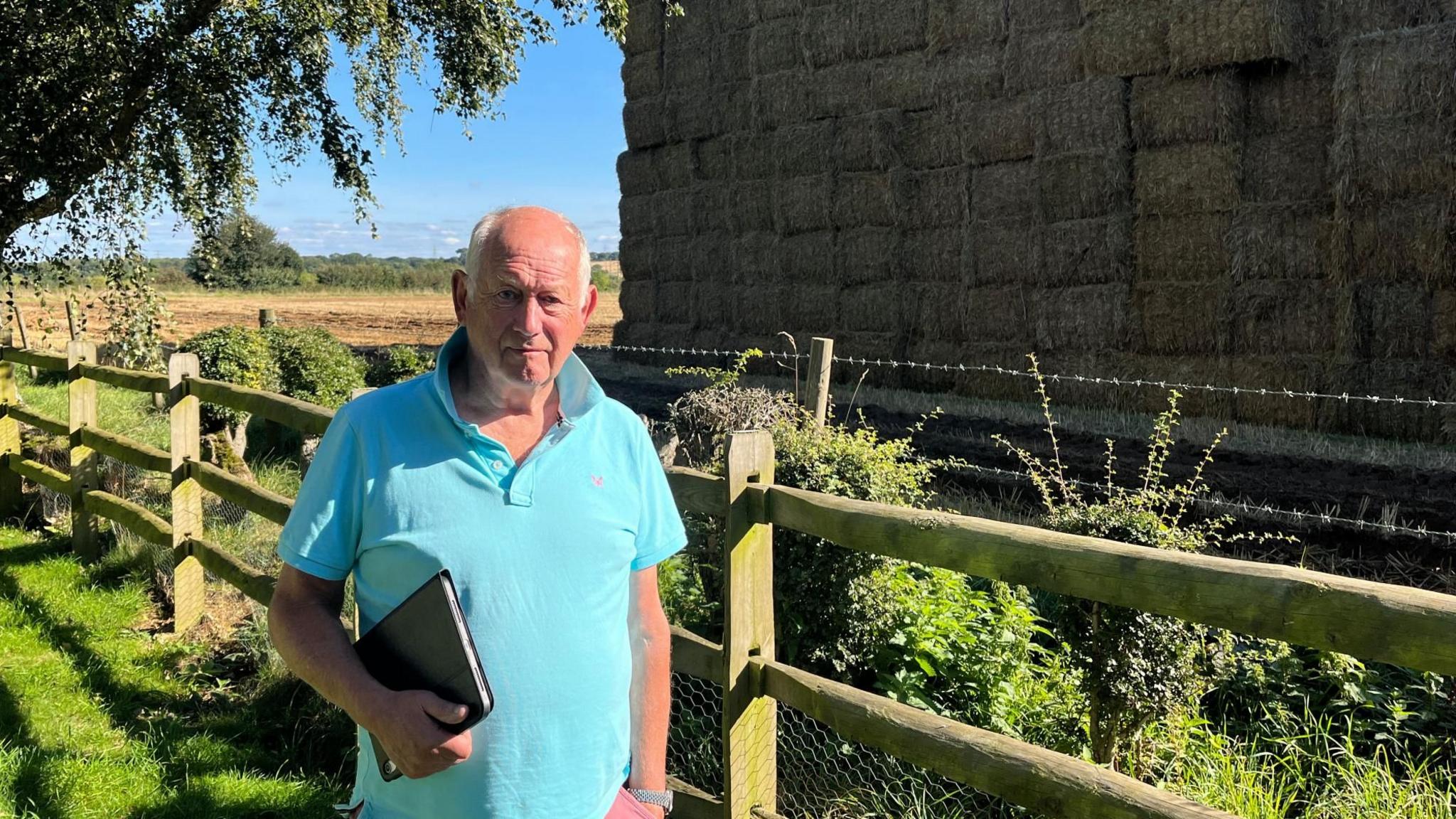 Cliff Nye is wearing a blue polo shirt and standing next to the giant haystack and close to a tree and a boundary fence.