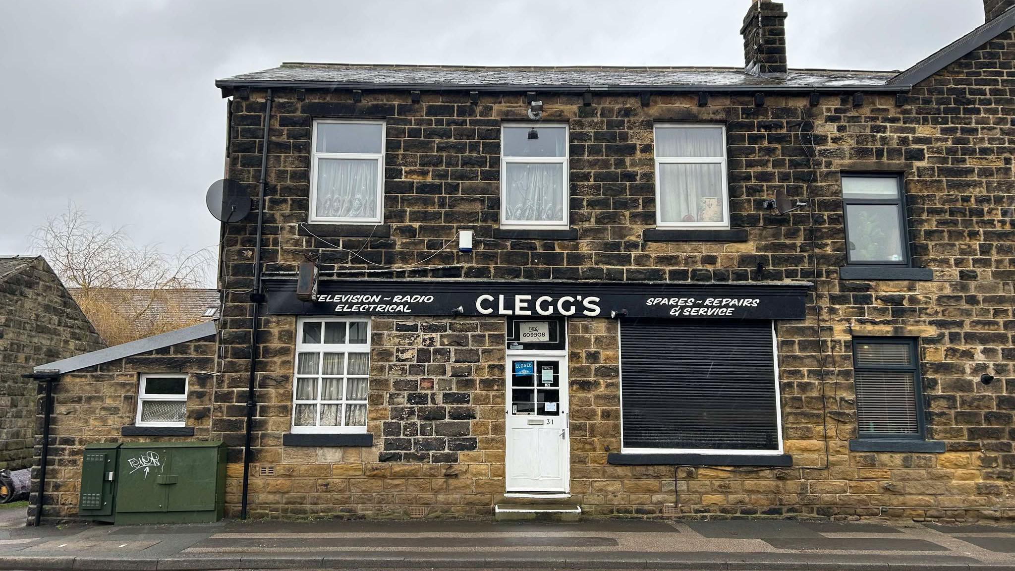 An electrical shop in a Yorkshire stone building with a sign which reads Clegg's. There are net curtains at the window and the stone is black from soot. 