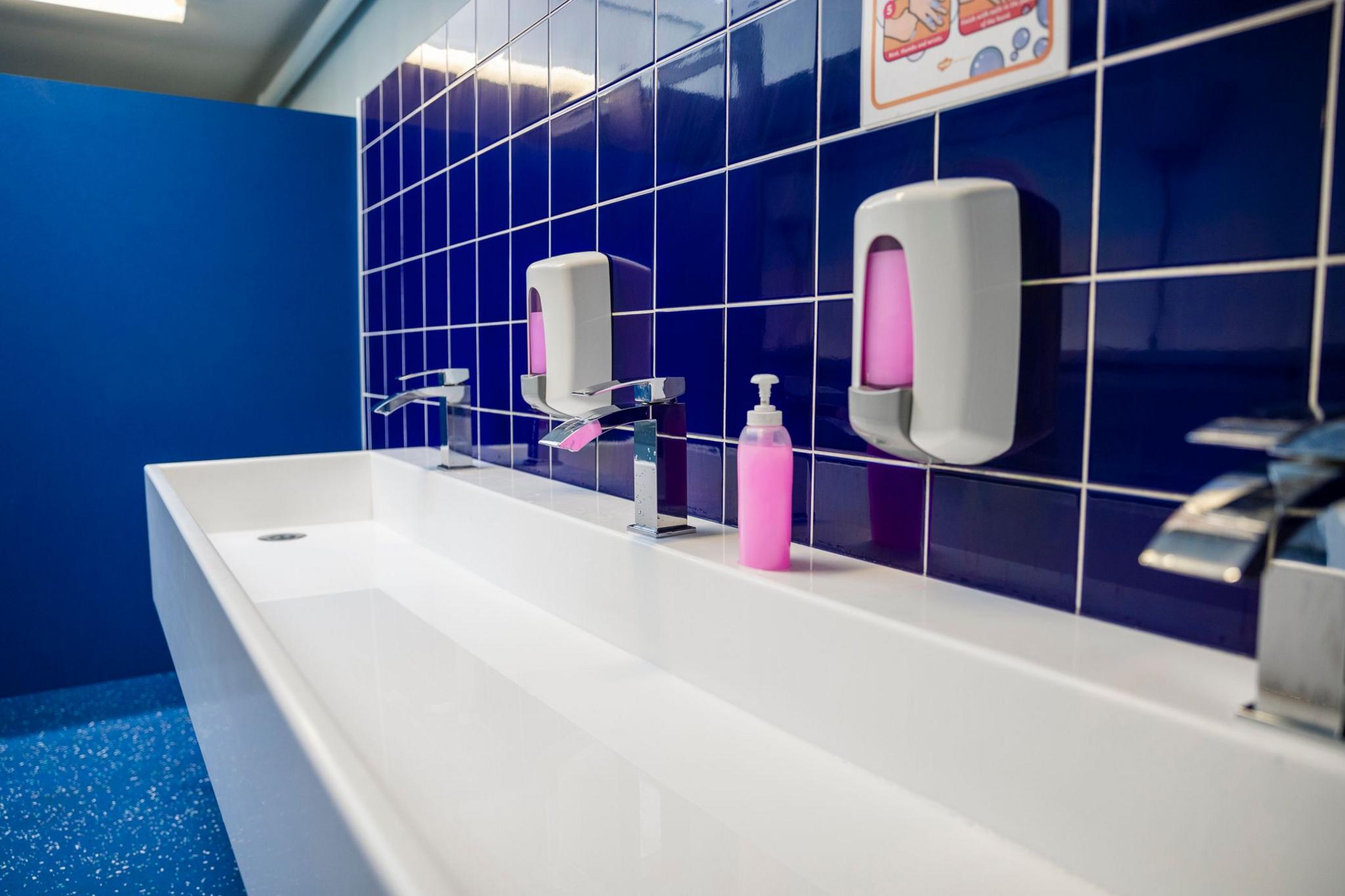 A close-up shot of a sink in a school bathroom featuring blue tiles with white grout and pink hand soapo.