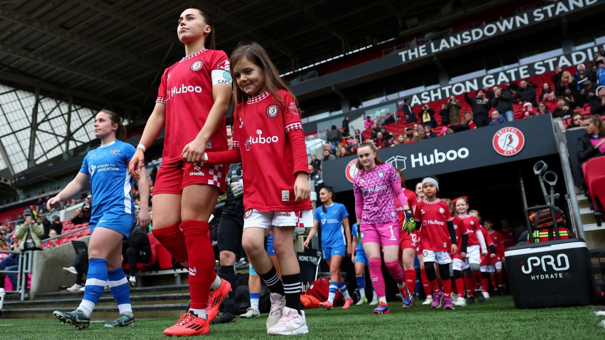 Players from Bristol City and Durham's women's walk out of the players' tunnel at Ashton Gate Stadium, some of the players hand in hand with young mascots. Bristol City players are in red and white and the Durham players are in their kit of blue and white