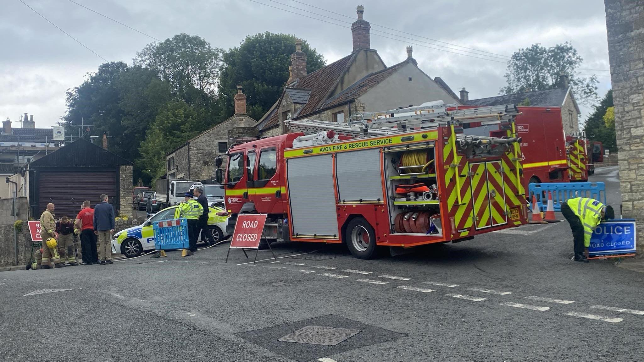 Fire engines and a police car on a street corner next to a red sign that says 'Road Closed'. A police officer is also placing a blue sign that says 'Police. Road closed' at the corner of the road.