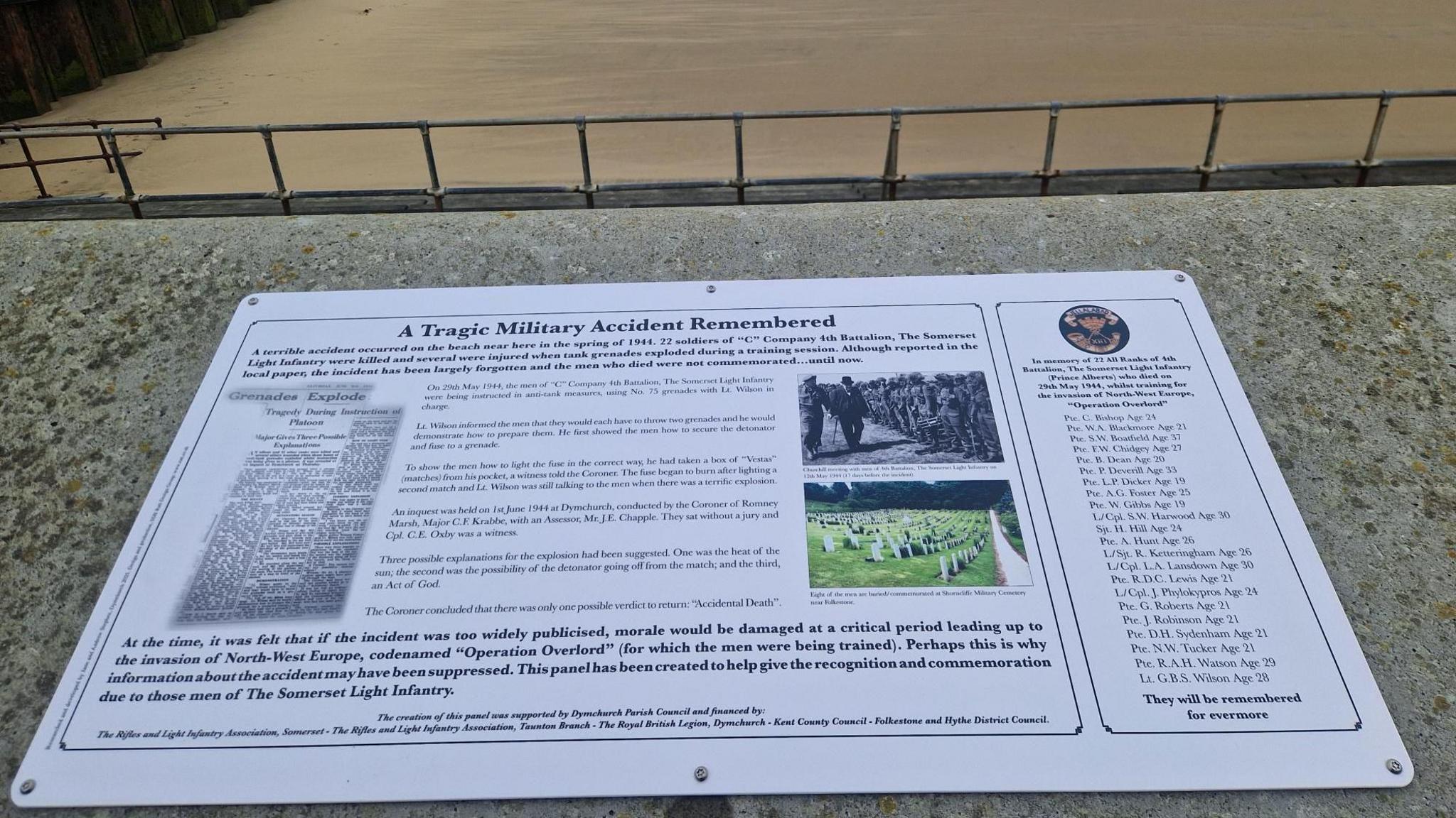 A large white plaque on top of a seawall, which details "A Tragic Military Accident" on 29th May 1944. The photo also shows part of the beach itself and metal railings