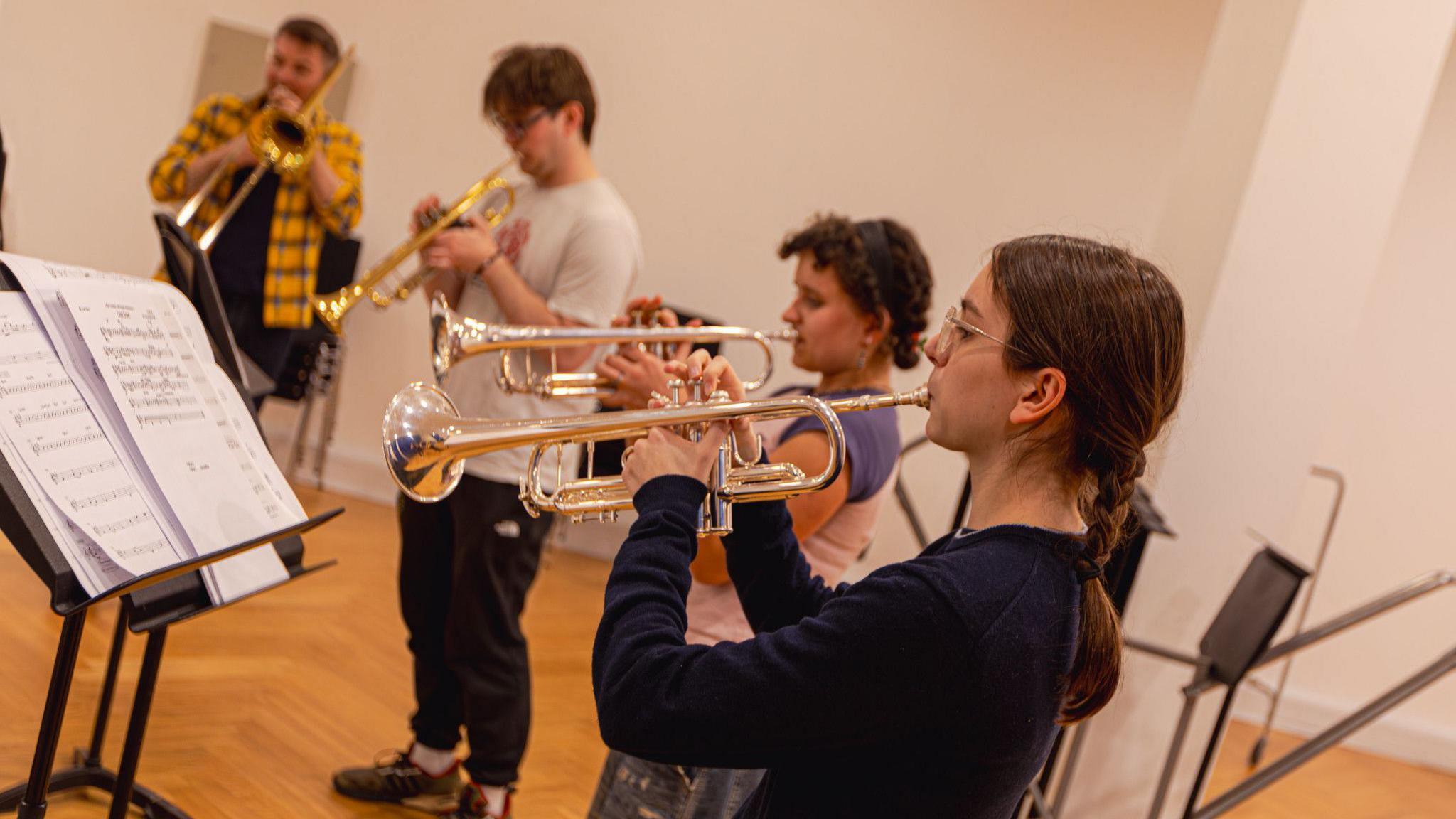 Four brass musicians playing in a room. Three people are playing trumpets, accompanied by a trombonist. They are standing in a semi-circle reading sheet music on traditional stands.