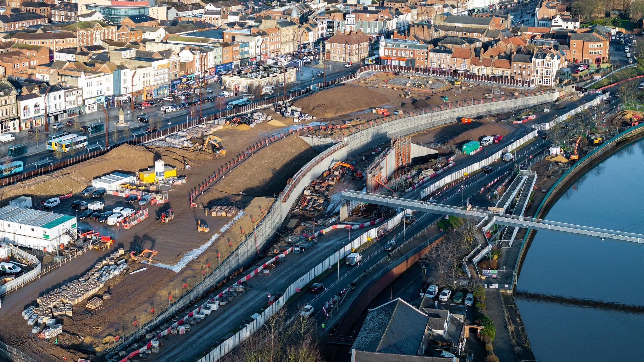 An aerial view of a narrow, modern white footbridge over the River Tees. The bend of the river can be seen to the right with the bridge disappearing out of shot. On the left, the river gives way to yellow building machinery on the bank. In the foreground is a building with a tiled roof behind which a line of cars are parked. A road along the bank has tall silver fencing on one side and short red and white barriers on the other. There is a large expanse of flattened earth between the road and the buildings of Stockton.