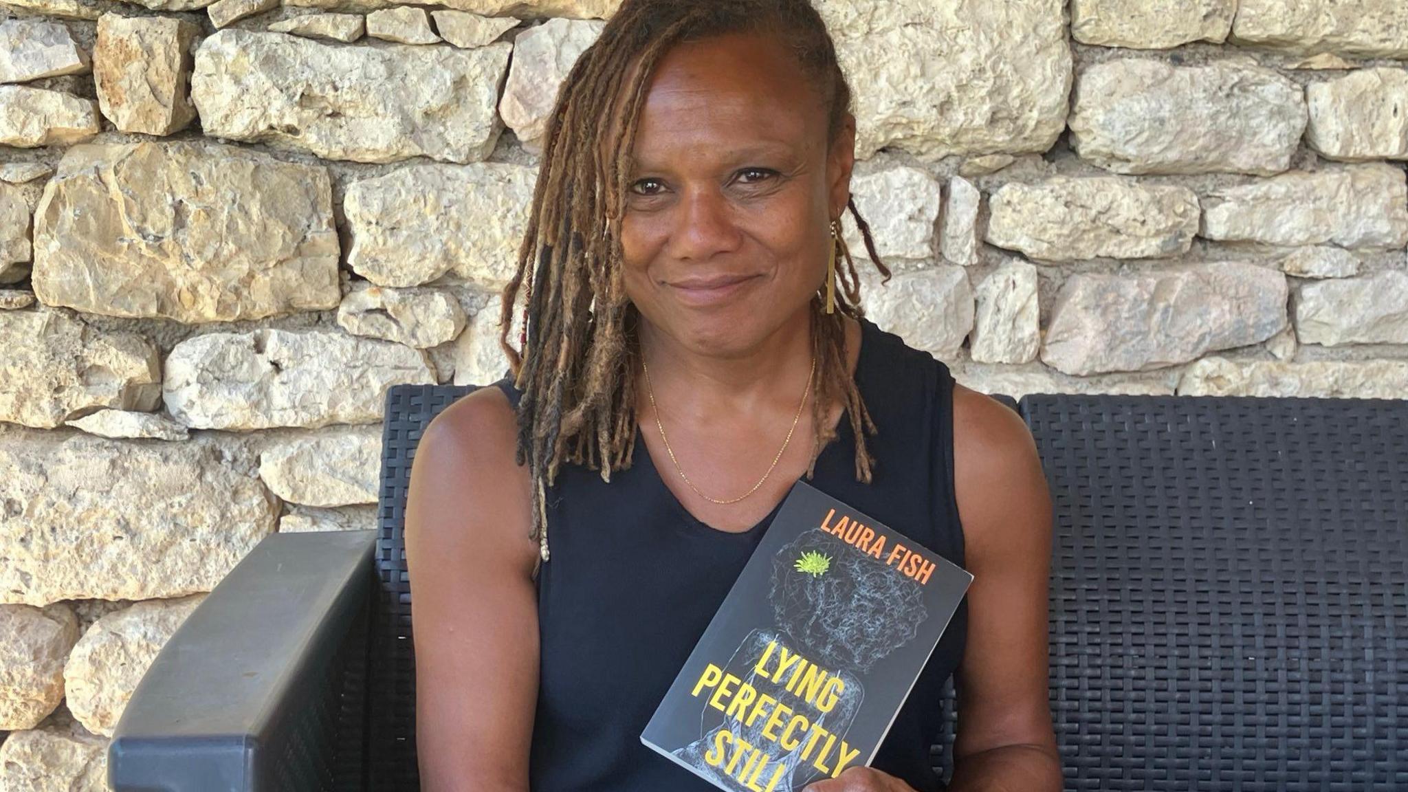 Laura Fish sits on a grey wicker garden chair, smiling and holding a book entitled in yellow lettering Lying Perfectly Still and with her name in orange lettering as author. She is wearing a dark vest top and has her brown hair in lots of thin braids. 