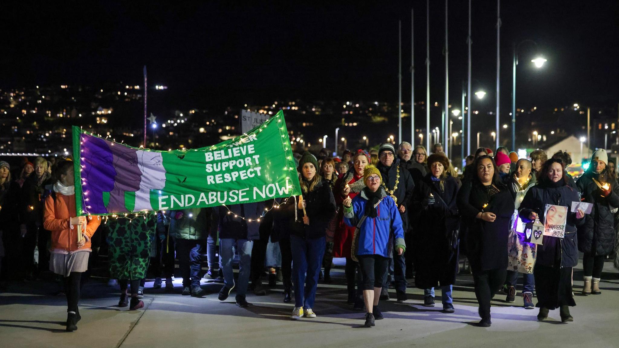 The photo shows people marching down a promenade in Penzance at night. There is a green banner that says "end abuse now".