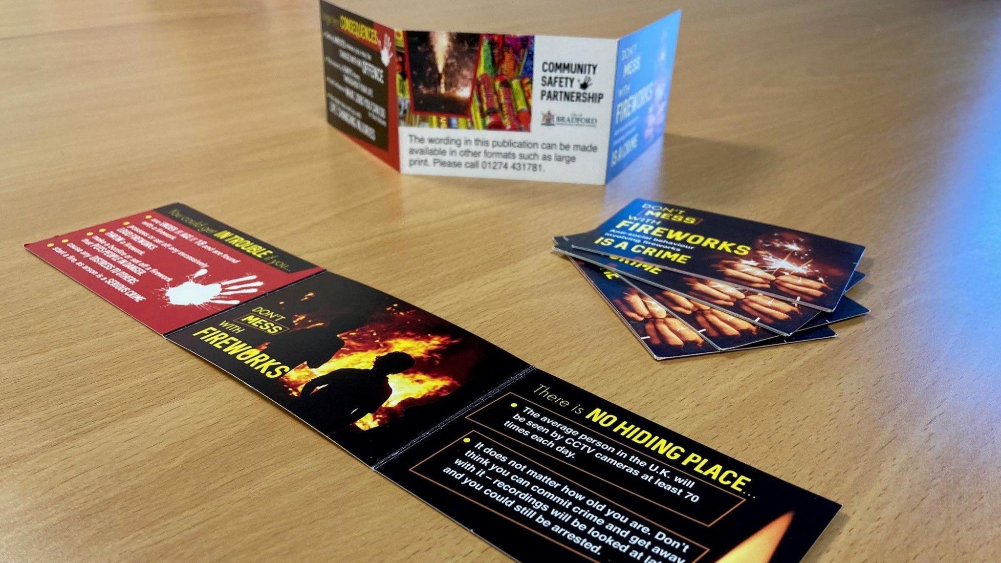 An unfolded leaflet laid out in the foreground on a brown table with four more folded up into bank card-sized shapes behind it and another leaflet in the background stood on its side.  