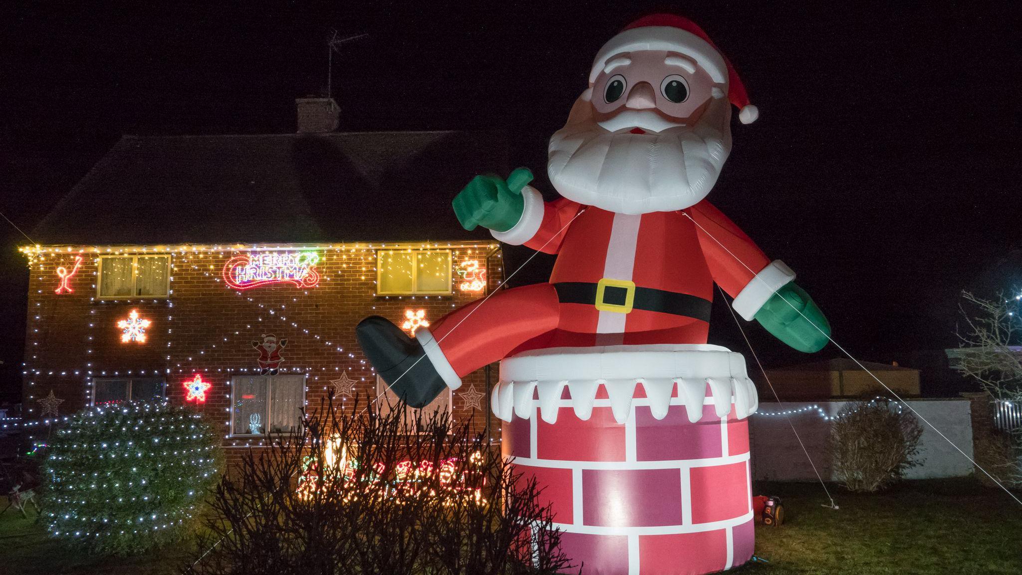 A giant blow up Santa which is the same height as the house it is next too is lit up. A house is covered in festive lights.