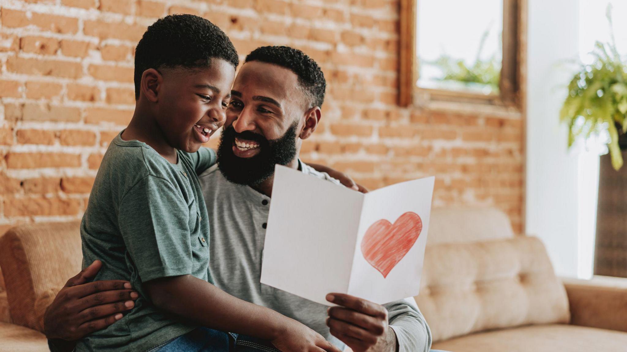 a child sitting on his dad's knee, the dad is holding a white card with a red heart drawn on it.