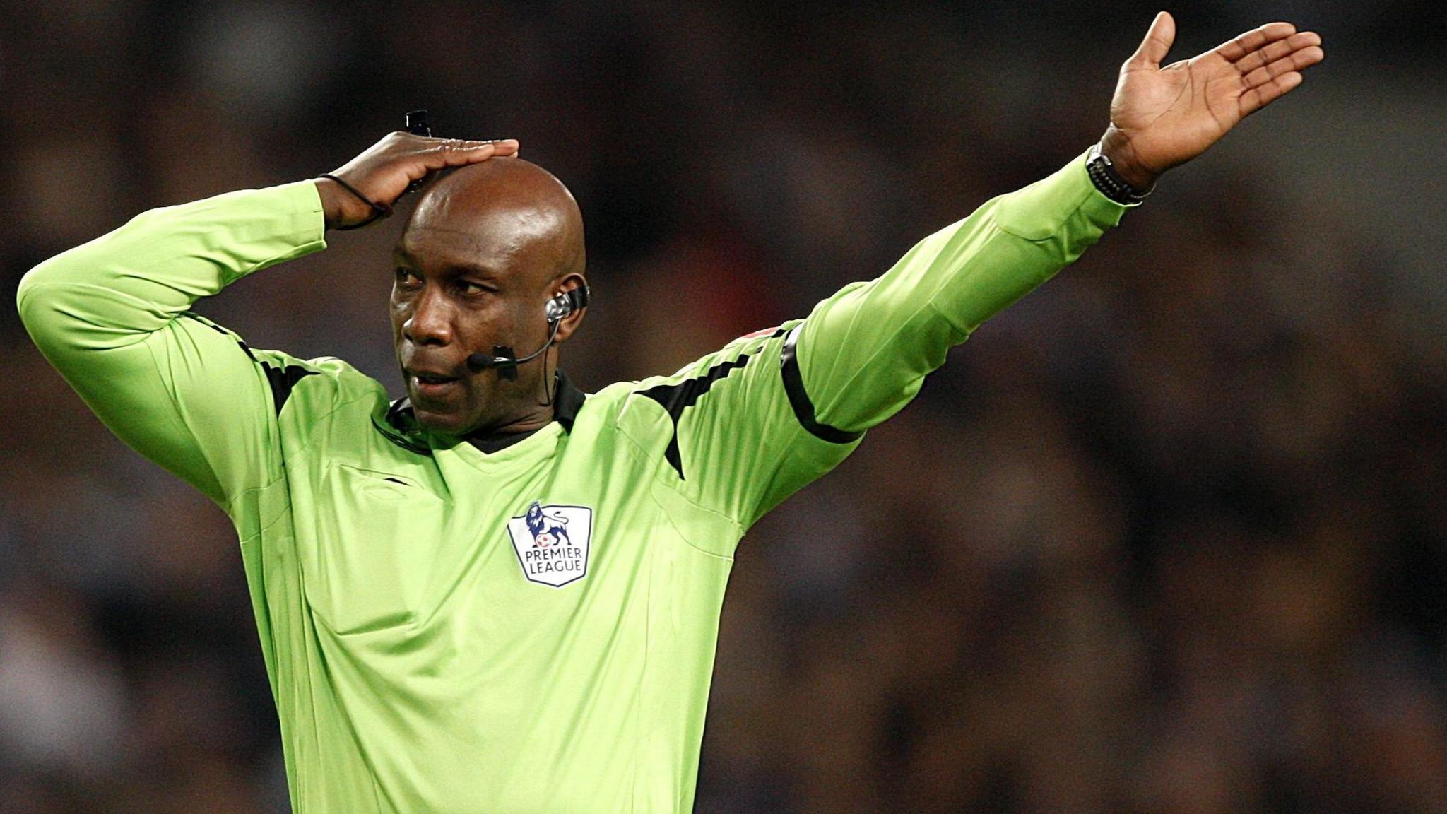 A man wearing a lime green referee's long-sleeved top with a "Premier League" badge on the chest. He is wearing a microphone and earpiece. He has one hand on his head and another outstretched in the air. He is looking away from the camera. A blurred background.