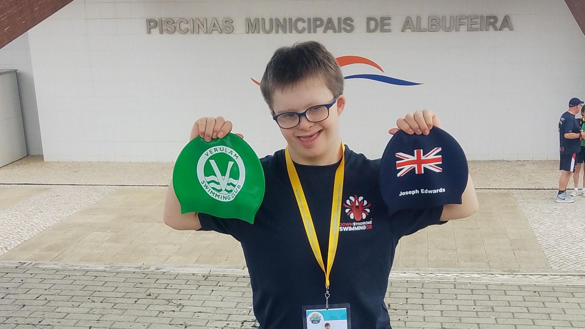 Joe, standing outside the entrance to a swimming pool in Portugal. He is holding up a green swimming cap and a blue one, he has a lanyard round his neck. He is wearing a blue T-shirt. There are two people in the distance to the right of the frame.