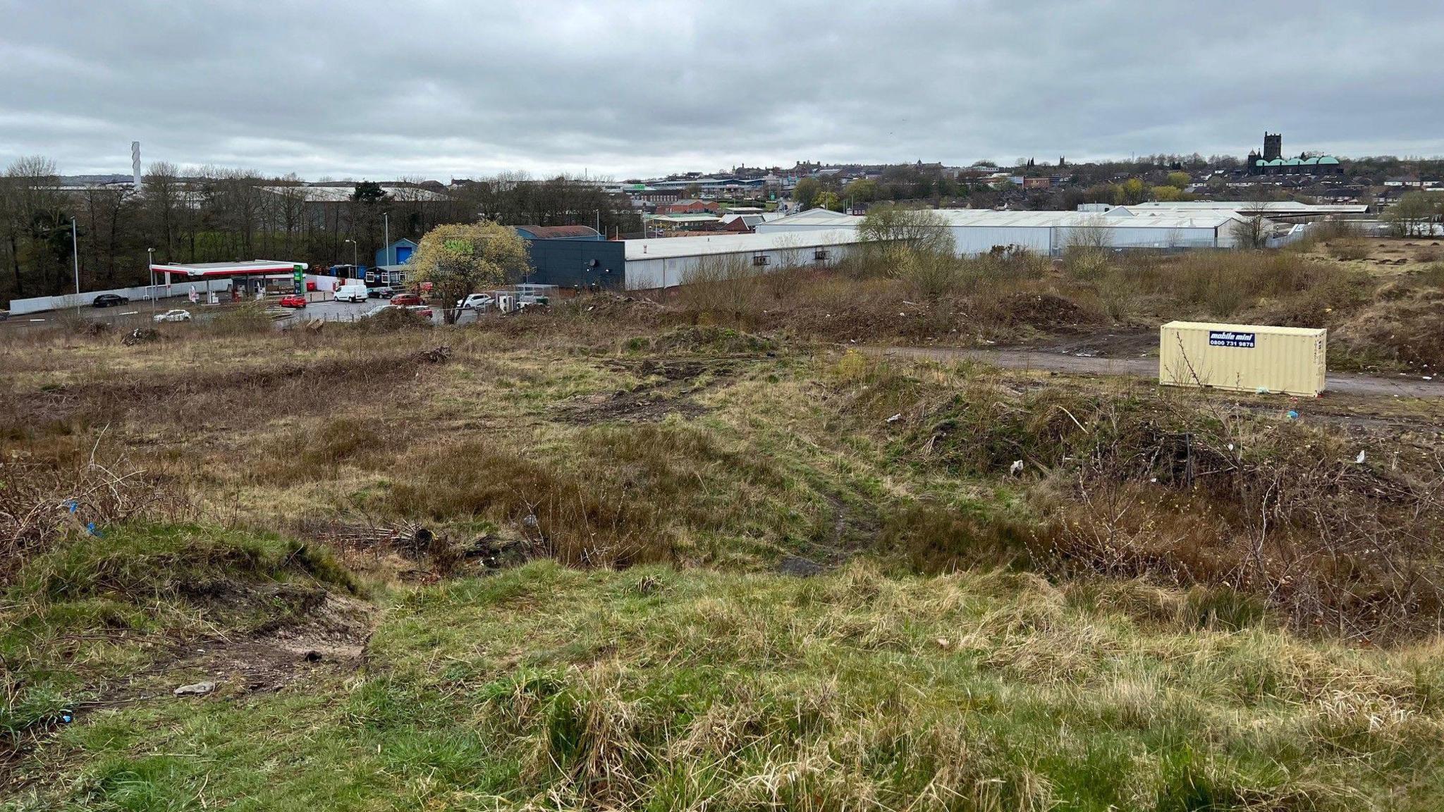 An area of land behind a set of industrial units with a petrol station and Tunstall town centre in the distance.