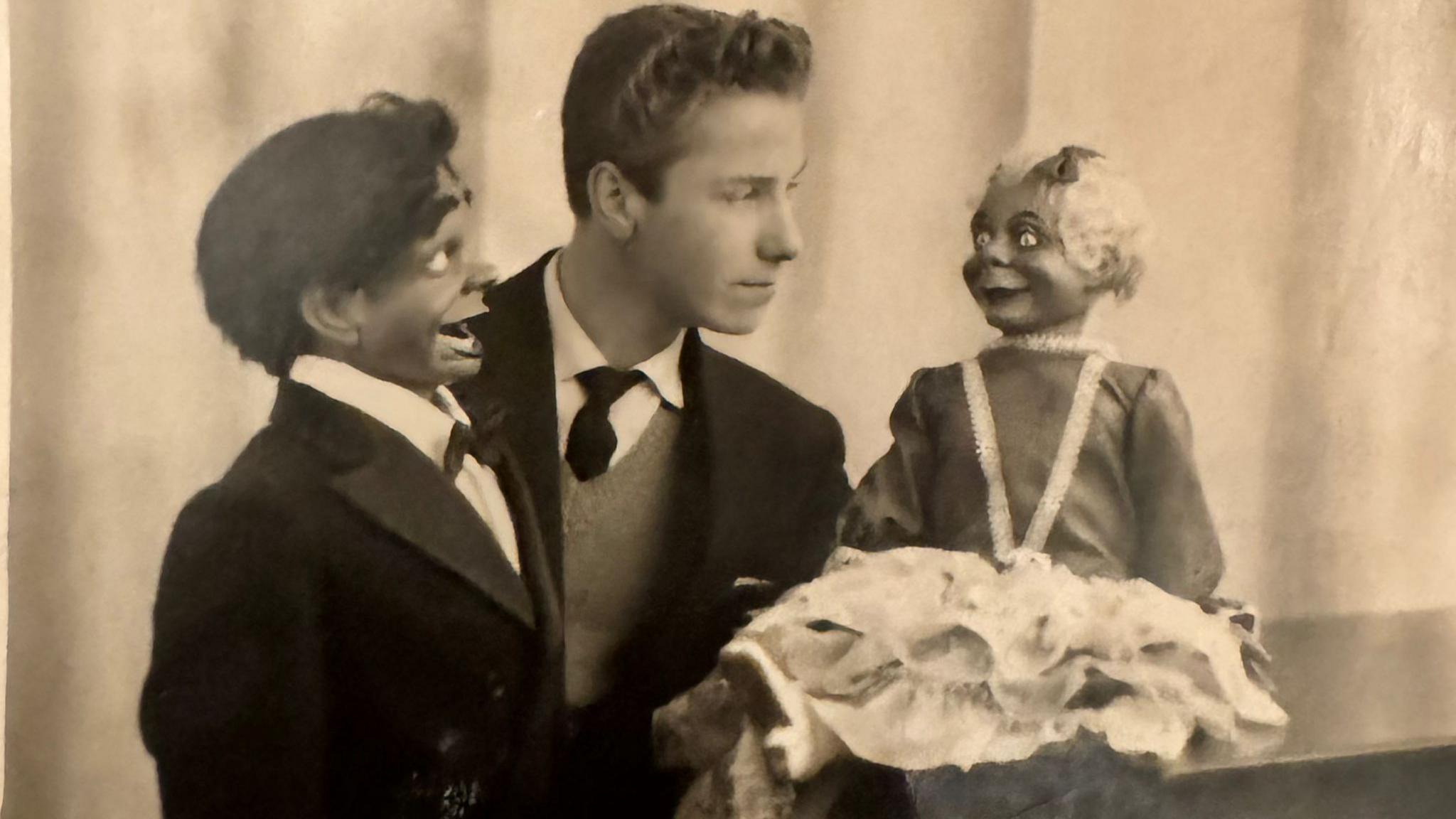 Mike Dennett in a black and white publicity photo taken when he was a young man. Mr Dennett is wearing a dark coloured suit jacket, a v neck jumper, white shirt and dark coloured tie. He is pictured with a male ventriloquist's dummy with dark hair and wearing a double breasted suit and bow tie and a female ventriloquist's dummy with blonde hair and wearing a dress.  