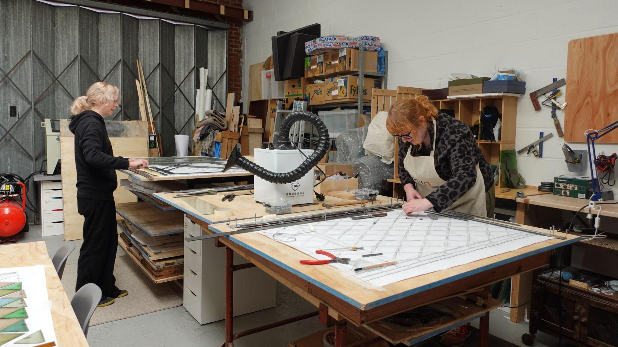 A woman with blonde hair and wearing a black top and trousers is stood at a workshop bench and working with a large pane of glass. On the other side of the bench a woman with red hair and wearing glasses, a black top and trousers and an apron is leaning over a large piece of paper and using tools.