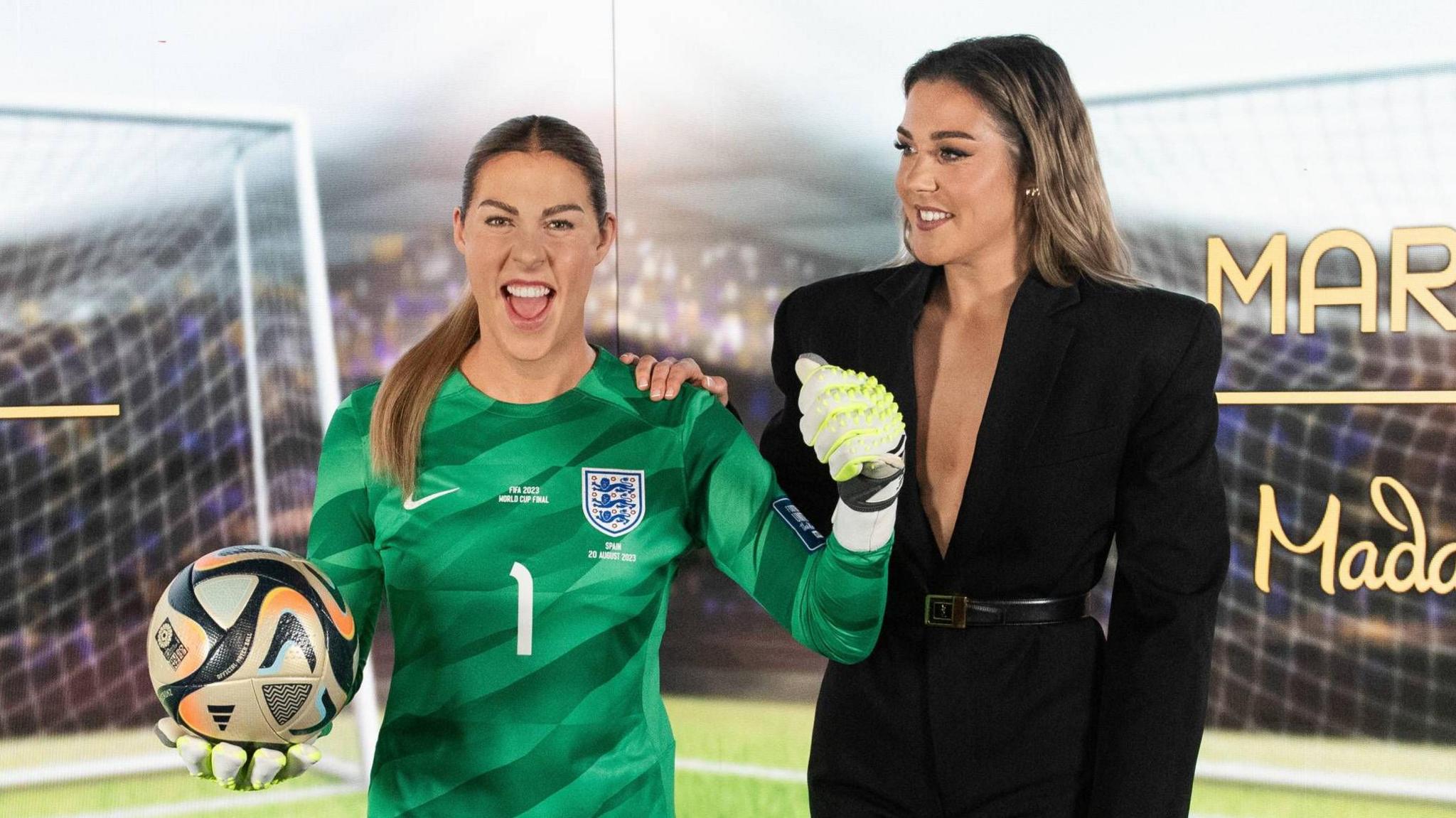 Mary Earps (right) wearing a black dress with long sleeves and padded shoulders. She is looking to her right and has her right hand on the shoulder of a waxwork in her likeness, which is modelled on her wearing an all-green England kit.