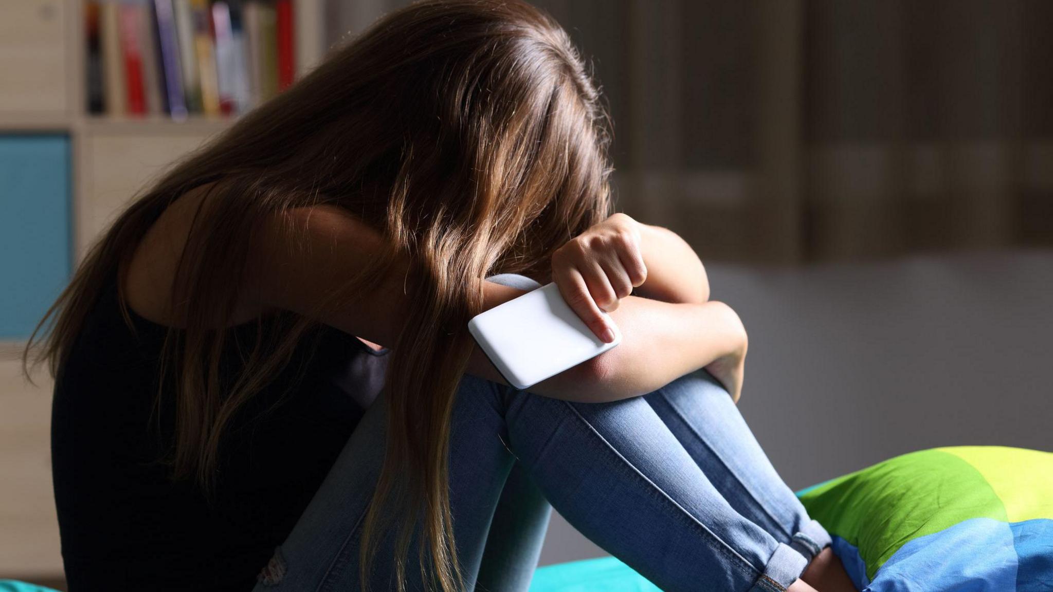 A young woman with long dark hair sits on a bed with her head in her arms. She is holding a white smartphone and is wearing a dark sleeveless top and light blue jeans.